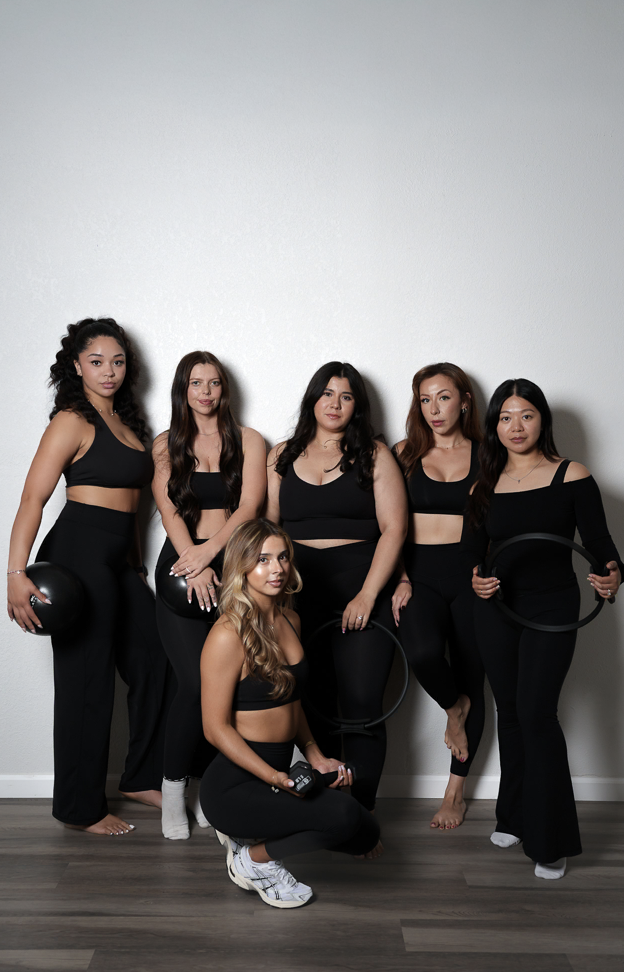 Six women in black athletic clothing posing indoors, with some holding fitness equipment like a medicine ball, yoga ring, and weights.