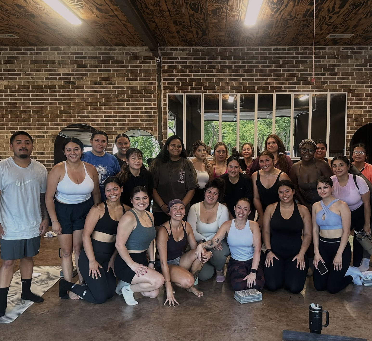 A group of people in workout or fitness clothing, smiling, posing together inside a gym or fitness studio with brick walls and large windows showing green trees outside.
