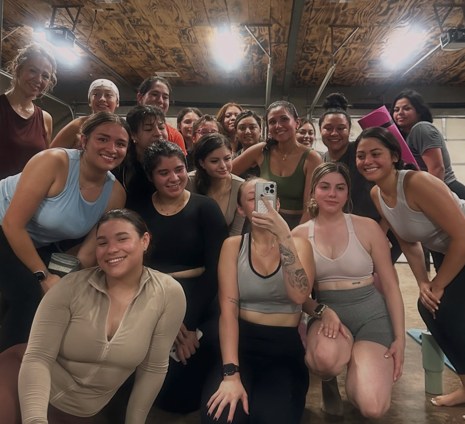 A group of women gathered in a fitness or yoga studio, taking a group selfie, with some women holding yoga mats and wearing workout attire.