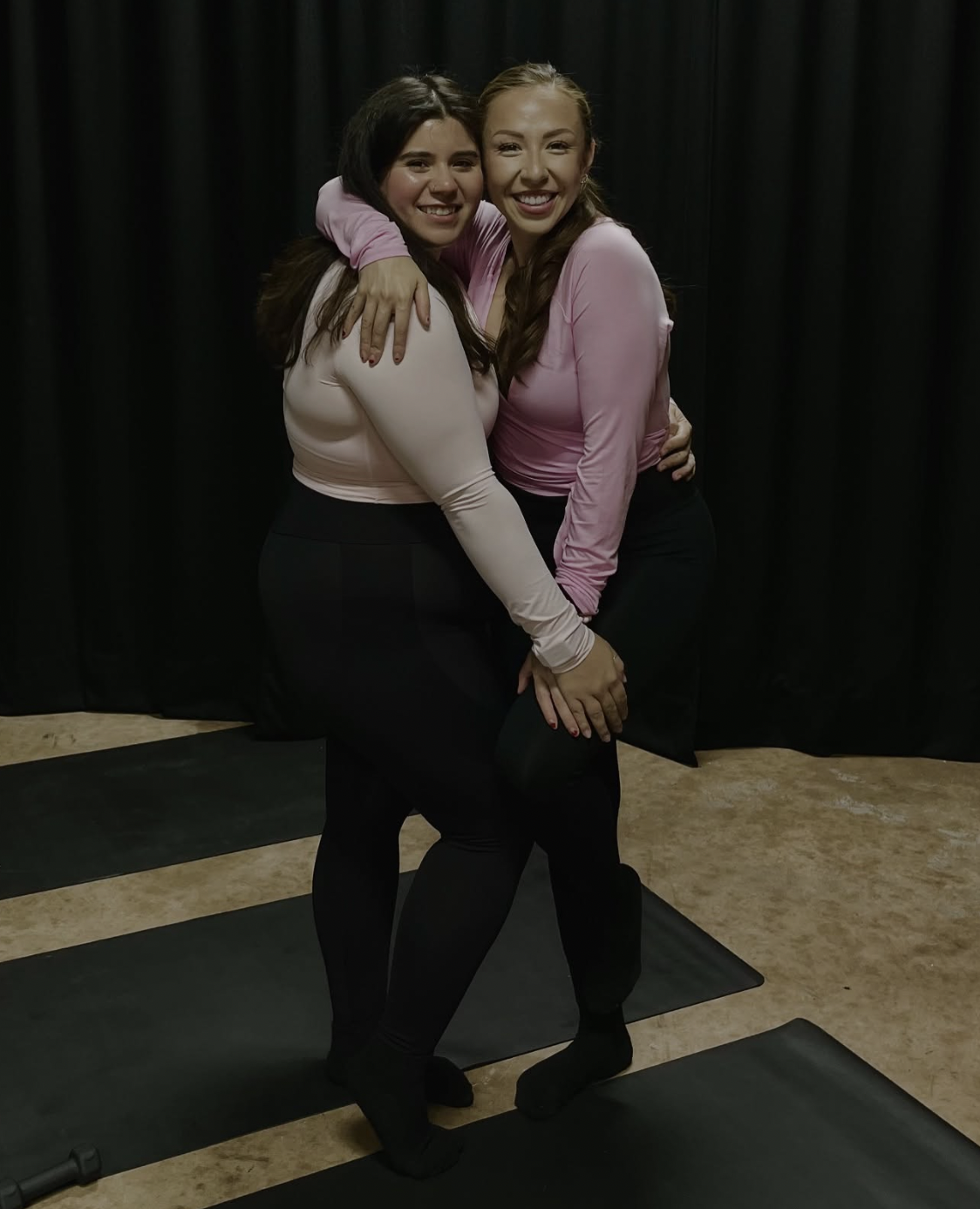Two women hugging and smiling, standing on black exercise mats in front of a black curtain.