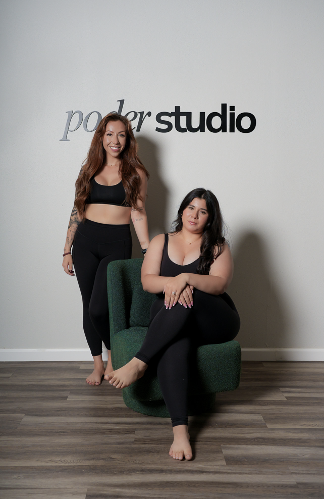 Two women in black athletic wear in a studio with a white wall that says 'power studio'. One women with long red hair is standing and smiling, and the other with long dark hair is sitting on a green chair with her legs crossed, looking at the camera.
