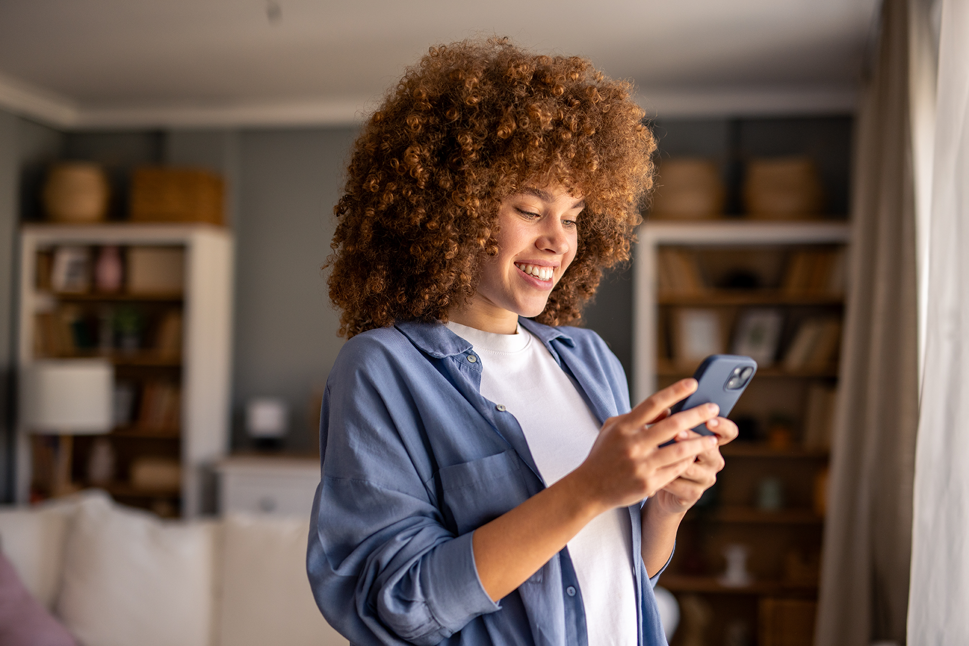 Smiling woman with curly hair looking at her smartphone in a room with bookshelves and a window with curtains.