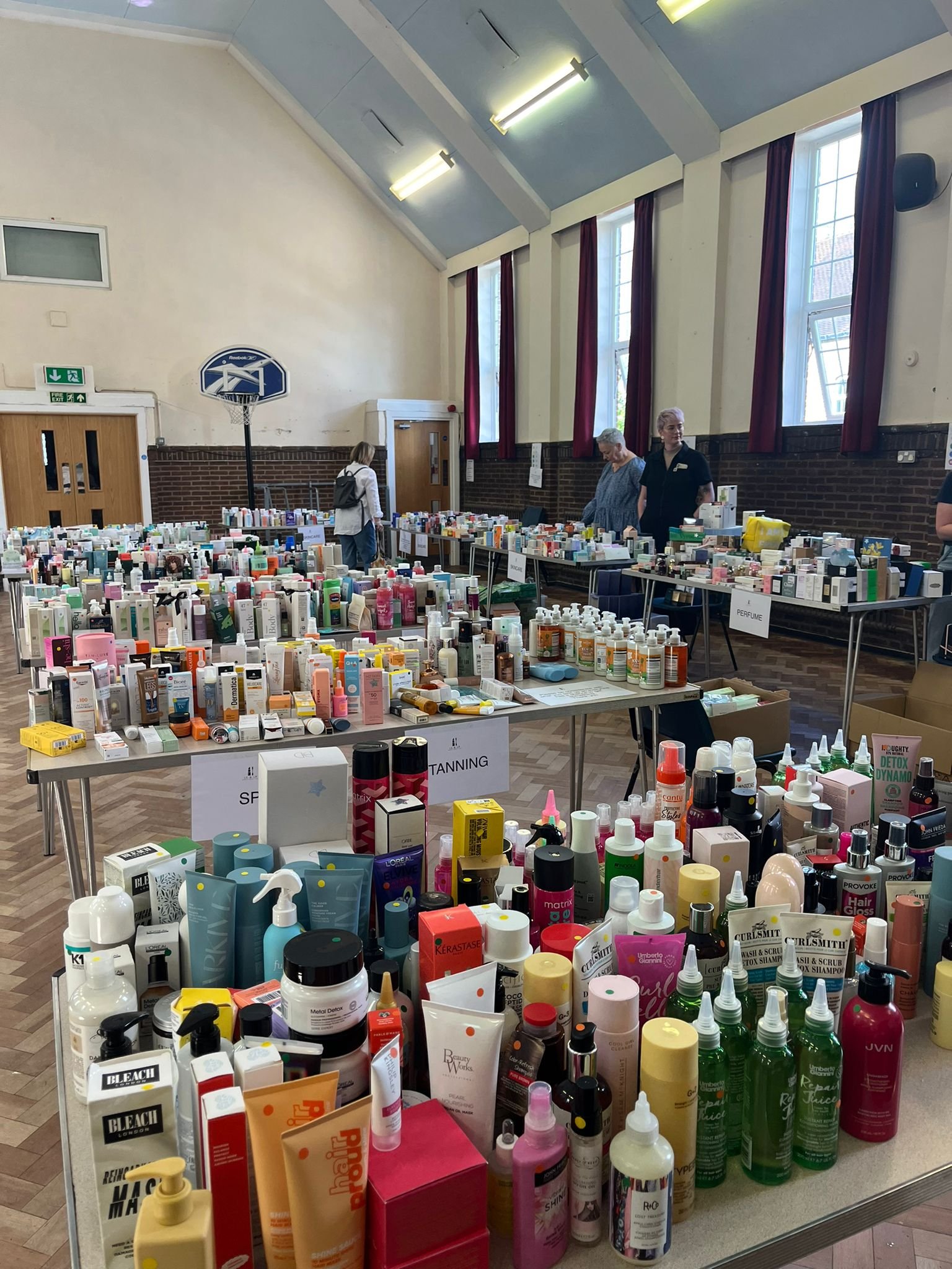 Tables filled with various beauty and skincare products at an indoor market or fair, with a basketball hoop and red curtains on windows in the background.