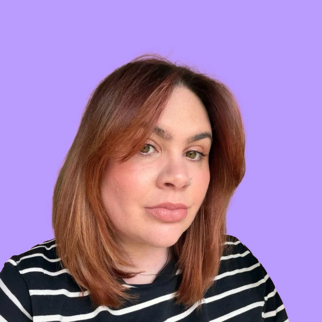 A woman with reddish-brown hair, green eyes, and a nose piercing, wearing a black and white striped shirt, looking at the camera against a purple background.