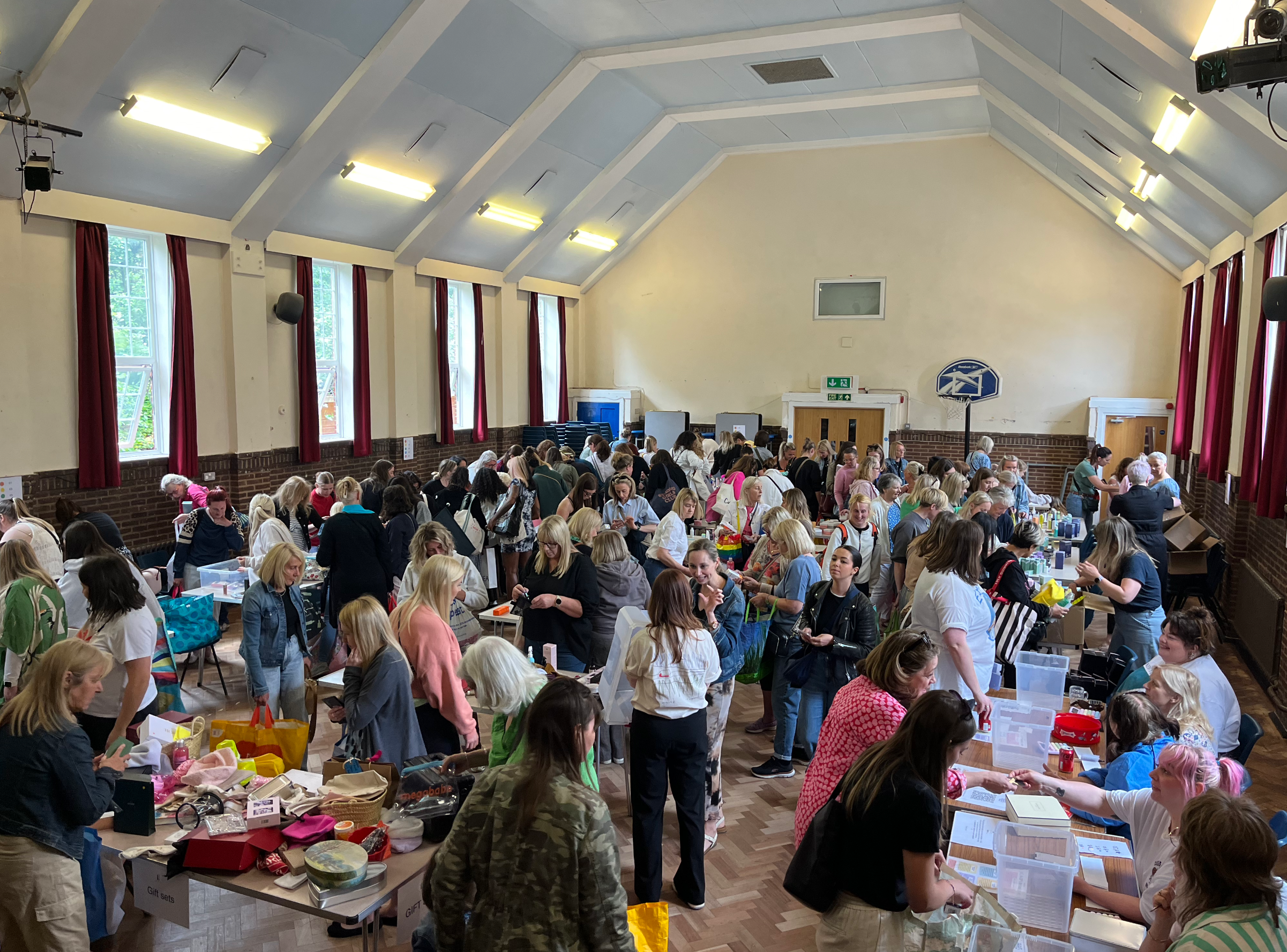 Large indoor market with numerous tables and people browsing and shopping, sunlight streaming through windows, red curtains, and a basketball hoop visible in the background.