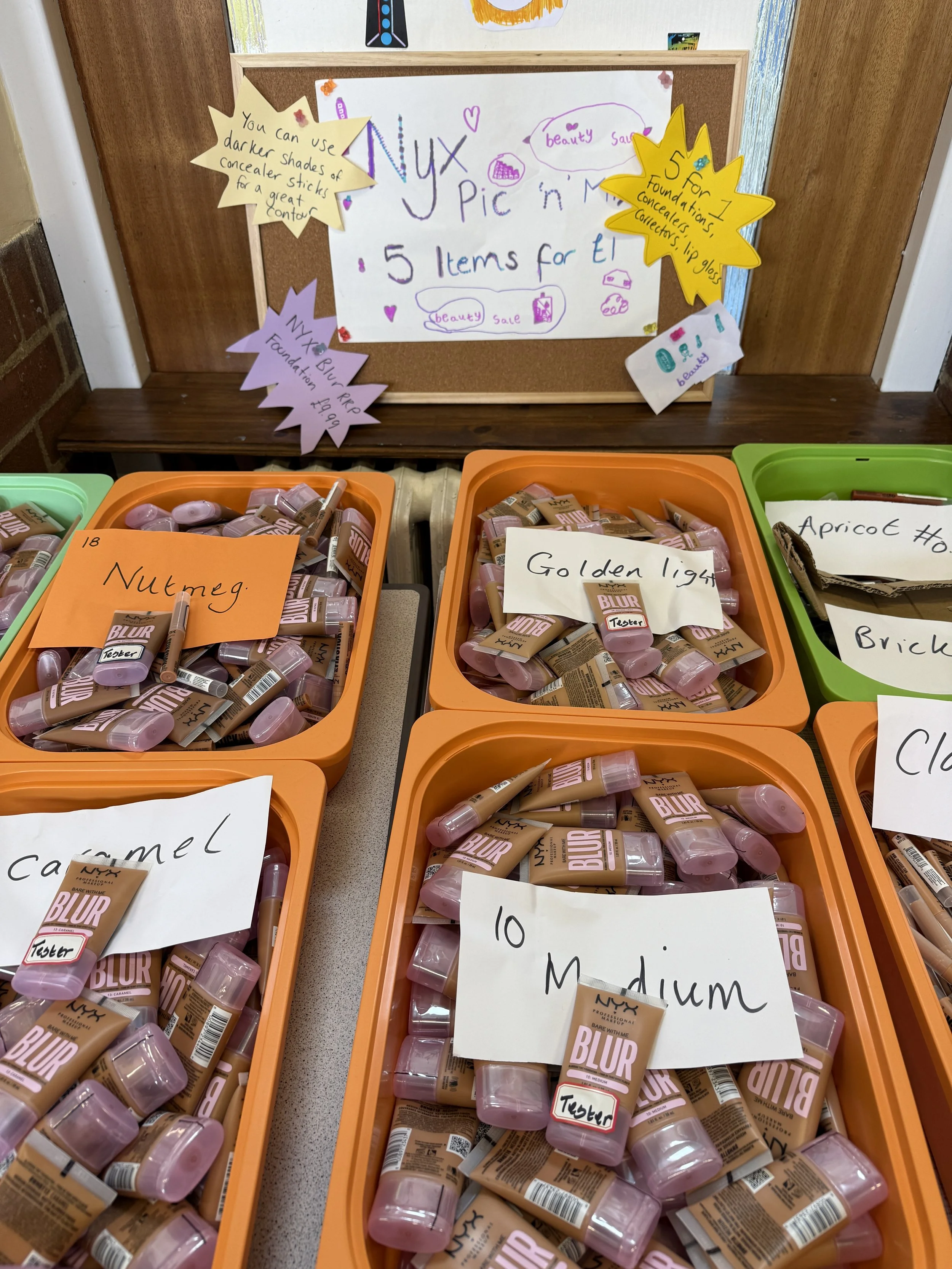 Display of five containers of NYX blush bottles labeled for sale, with handwritten signs indicating shades such as Nutmeg, Golden Light, Apricot, Caramel, and 10 Medium. A bulletin board behind the containers features colorful handwritten notes and s