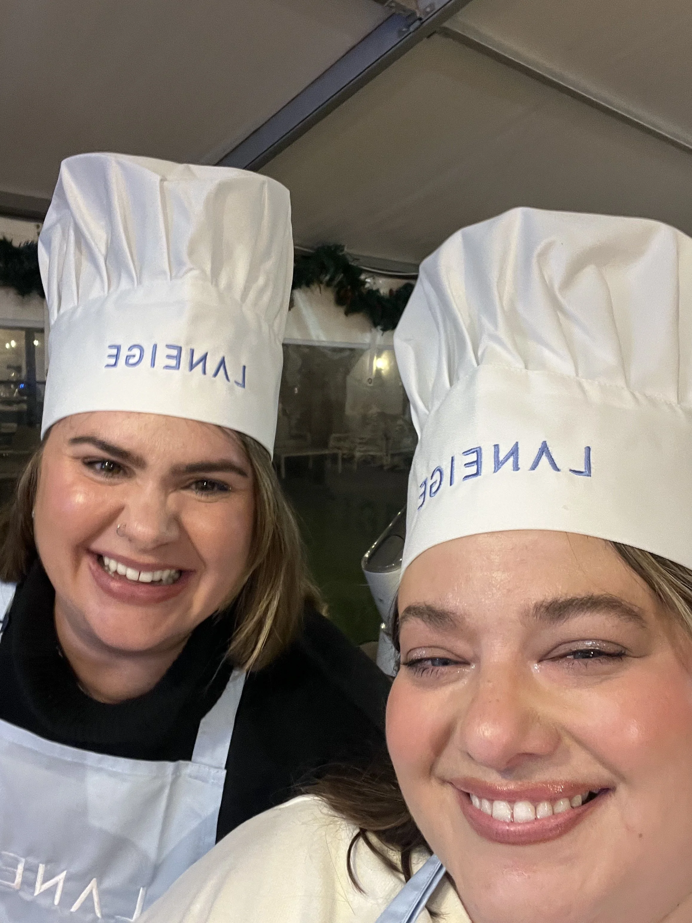 Two women smiling, wearing chef hats labeled 'LAVEIGE,' and aprons, in a kitchen setting.