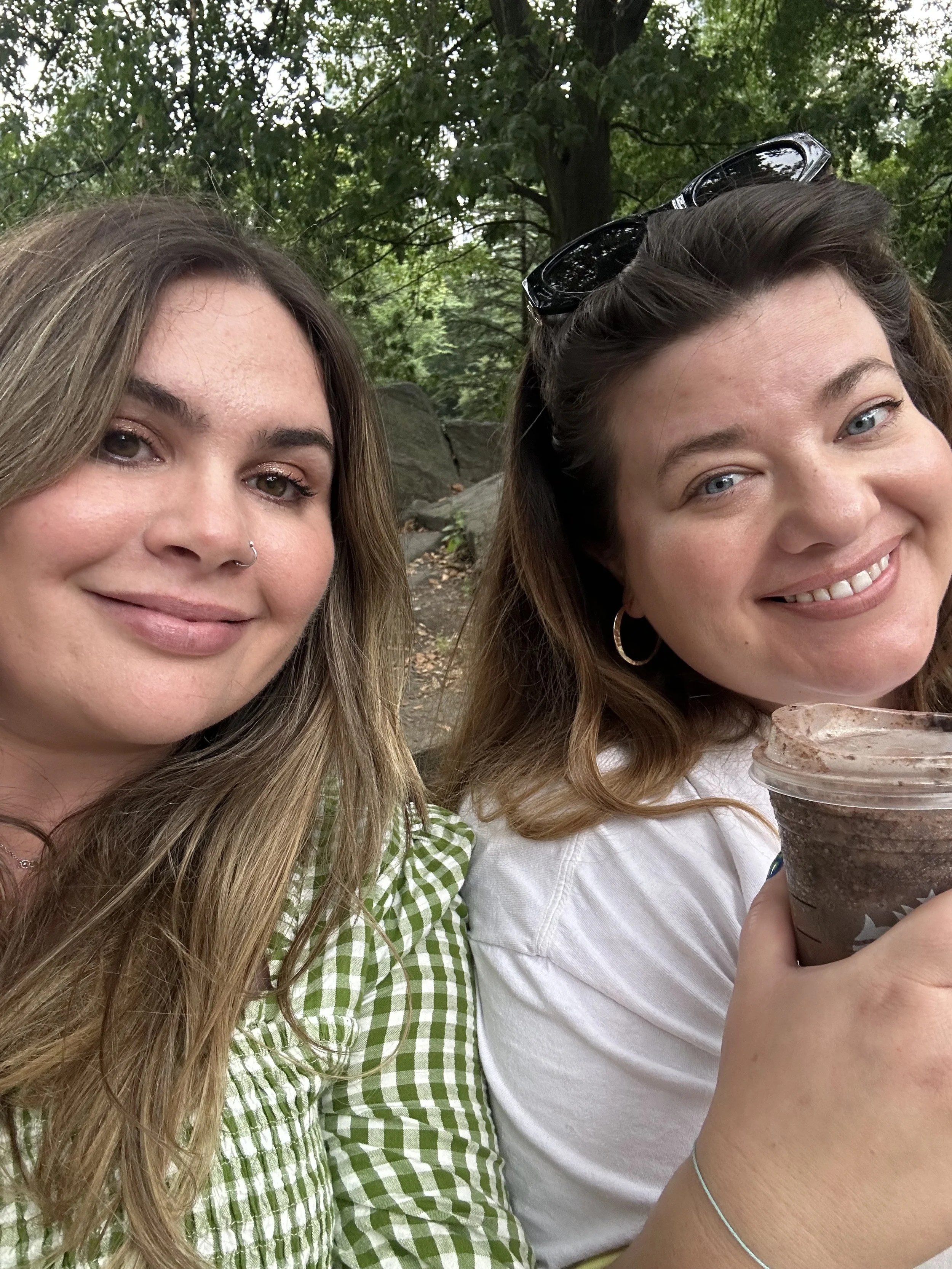 Two young women smiling outdoors on a cloudy day, with one holding a cup of iced coffee or chocolate drink.