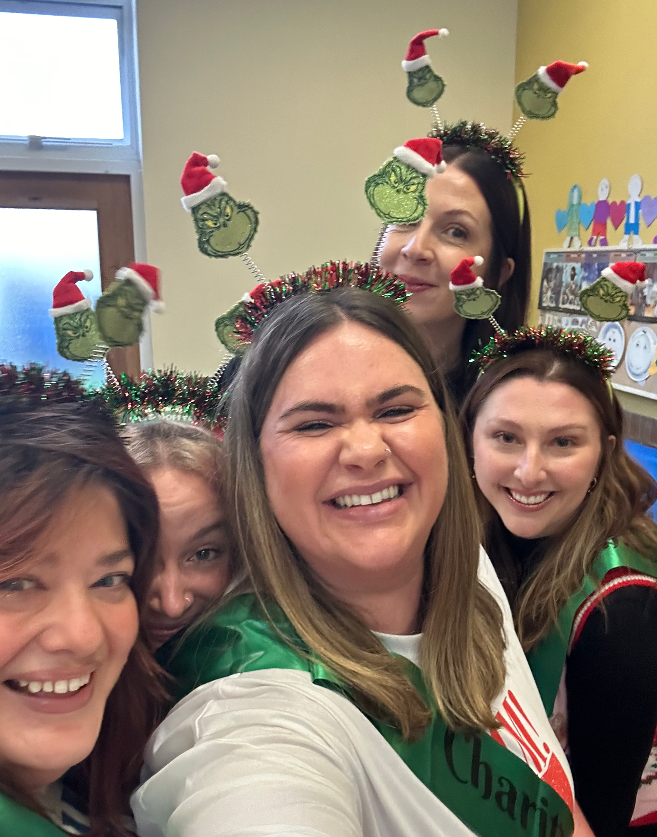 Five women at a holiday gathering, wearing Christmas-themed headbands with Dr. Seuss character hats, smiling and posing for a selfie in a decorated room.