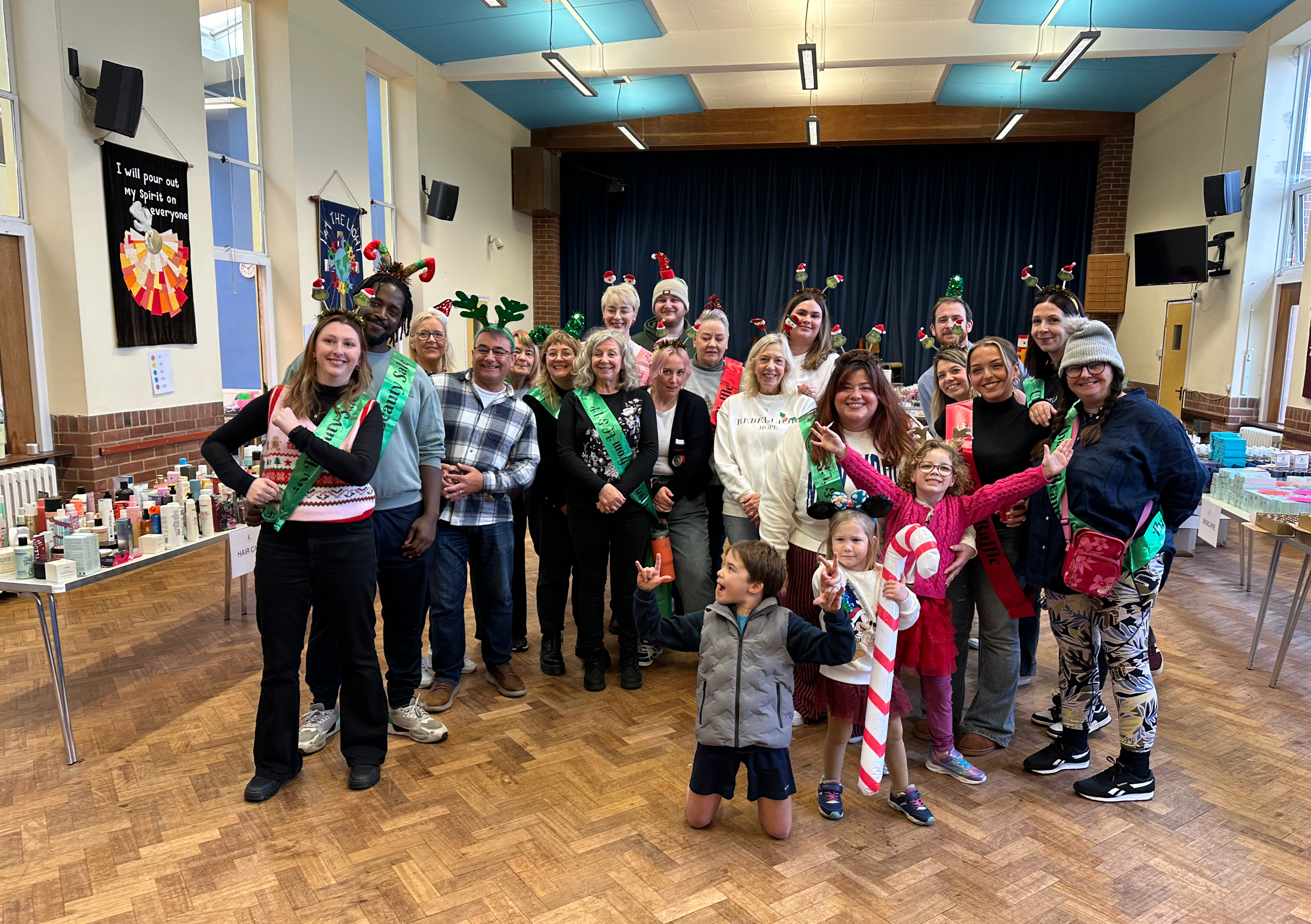 Group of children and adults celebrating Christmas in a decorated hall, some wearing holiday hats and reindeer antlers, with festive decorations and gifts in the background.