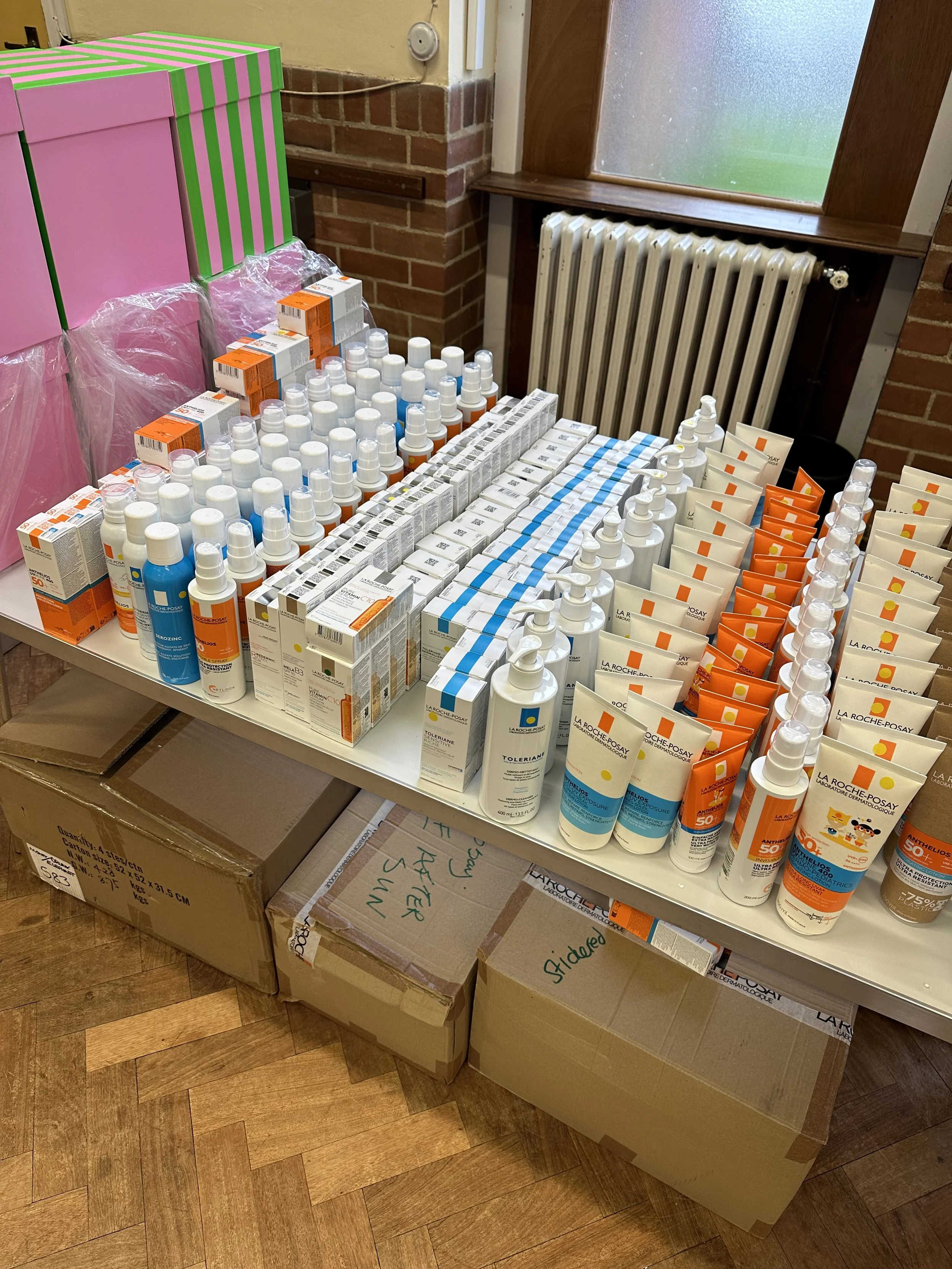 A display table with various bottles and boxes of skincare products, including sunscreens and lotions. The table is set against a brick wall with cardboard boxes underneath and a window with frosted glass in the background.