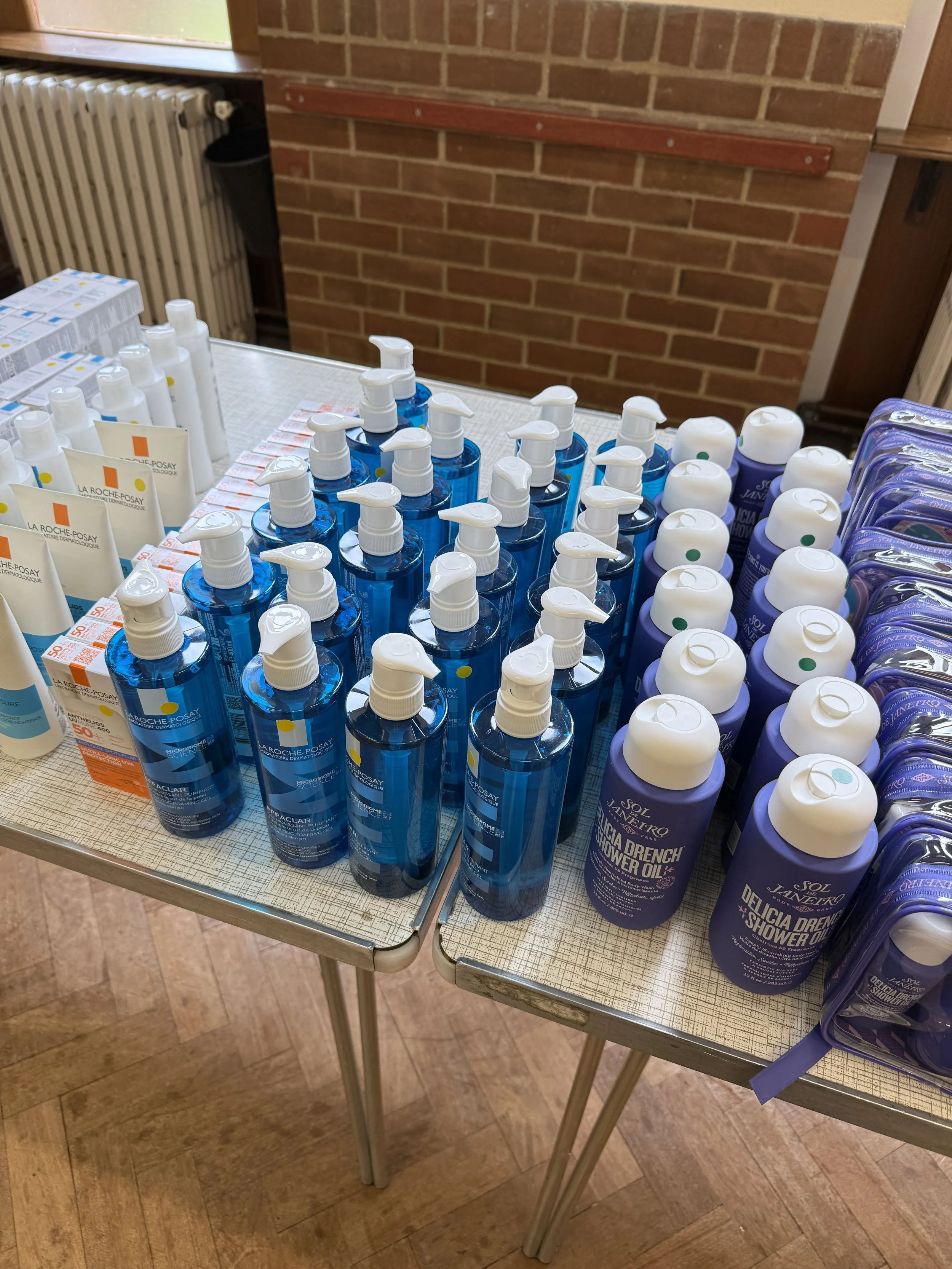 Display of personal care products on a table, including blue spray bottles, white bottles, and tubes, in a room with brick walls.