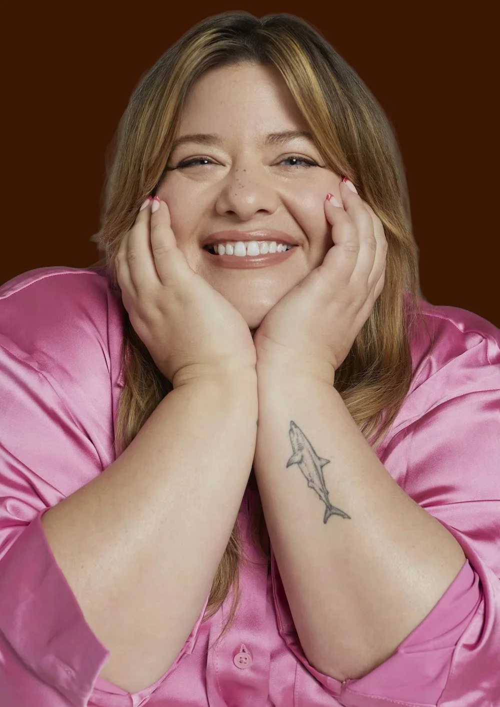 A smiling woman with long brown hair, wearing a pink satin shirt, with her hands on her face, and a shark tattoo on her left forearm, against a dark background.