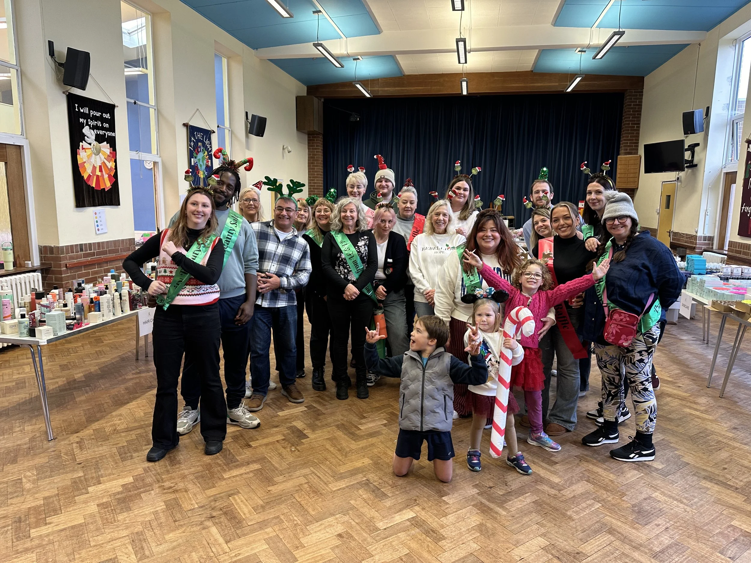 Group of people dressed in festive holiday attire, including Christmas headbands, antlers, and Santa hats, gathered in a decorated event hall with tables of products and holiday decorations.