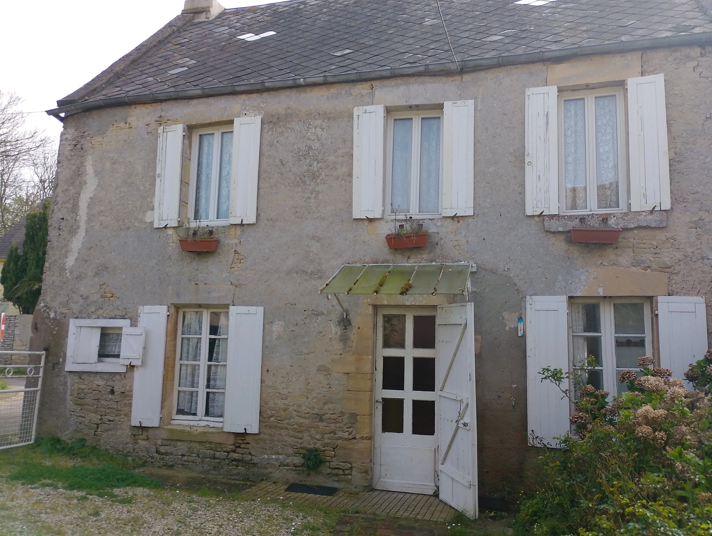 Old stone house with white shutters on windows, a white door, and small flower boxes beneath the windows. The house has a worn, weathered look, with a moss-covered glass awning above the door and some plants in front.