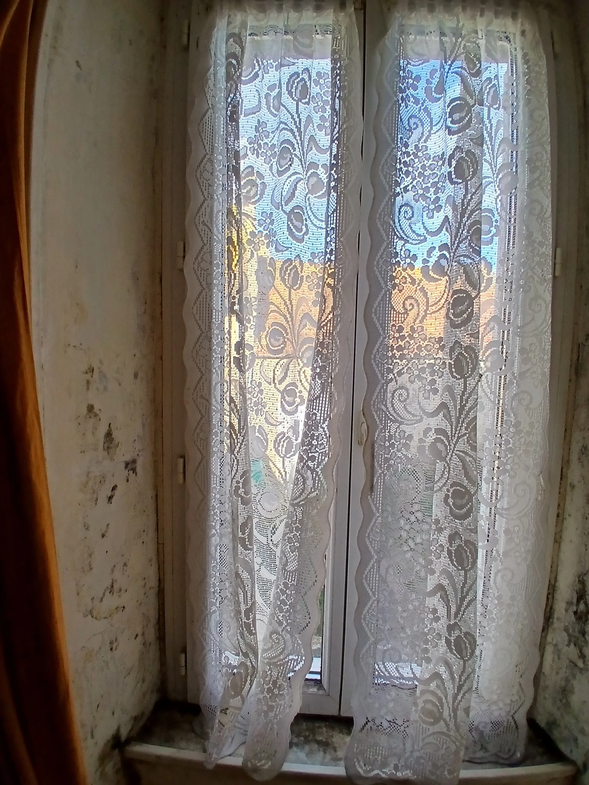 A window with sheer, patterned white lace curtains, partially open to reveal an outdoor scene with buildings and a sunset sky.