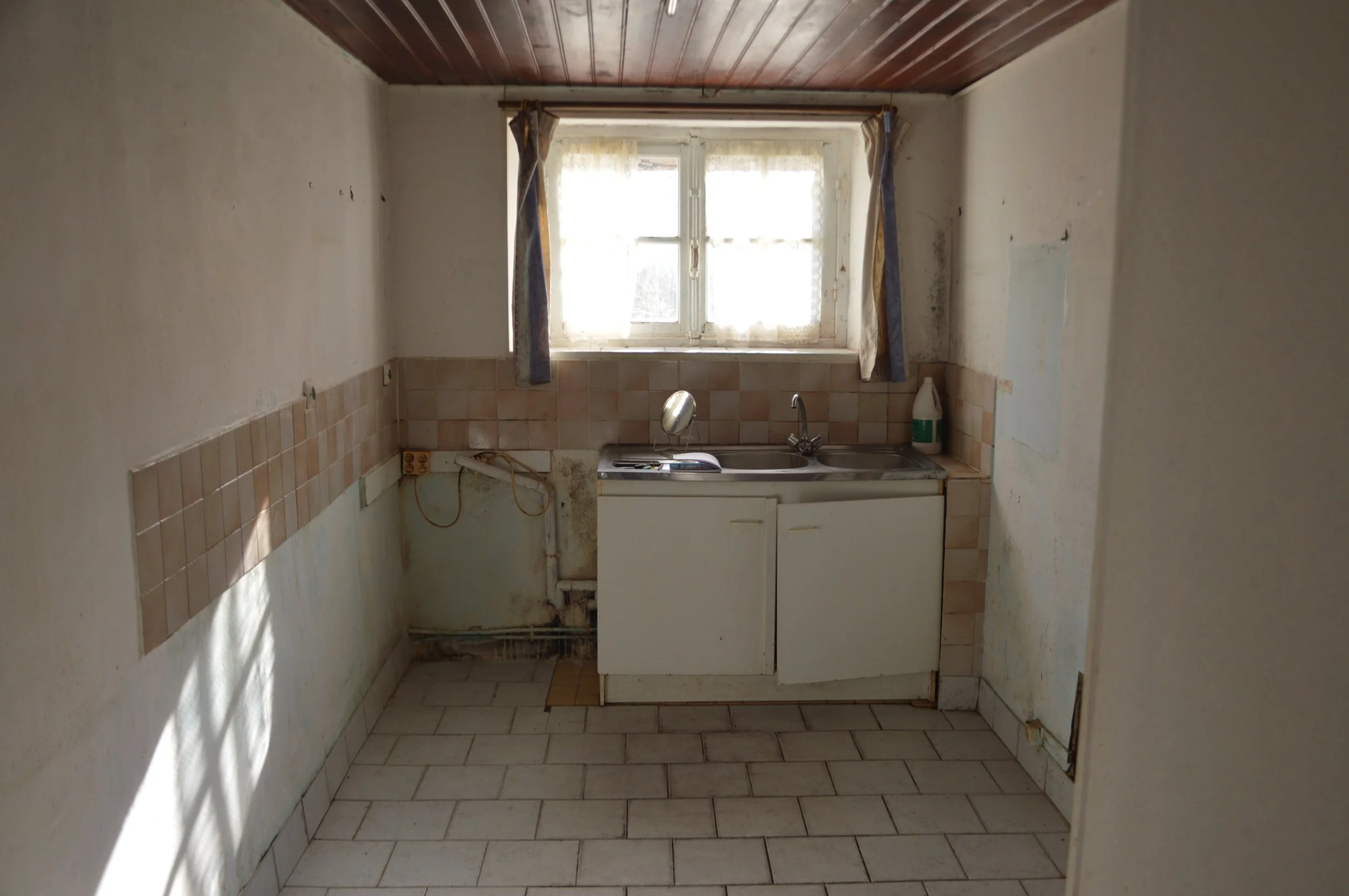 Old, empty kitchen with a small sink, a window with curtains, and peeling walls.