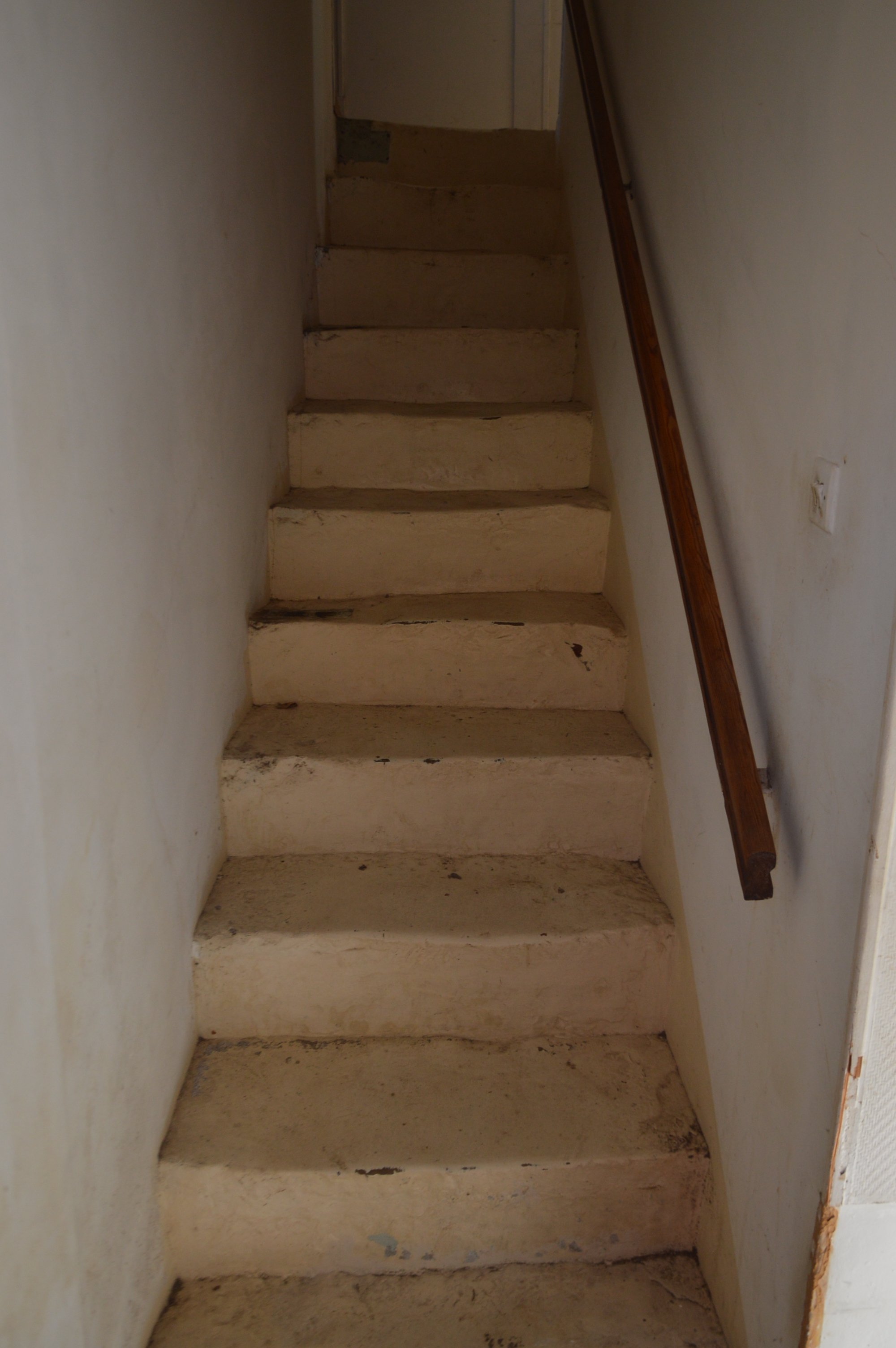 Old, uneven concrete stairs with a wooden handrail on the right side, leading up to an upper level in a narrow, dimly lit indoor space.