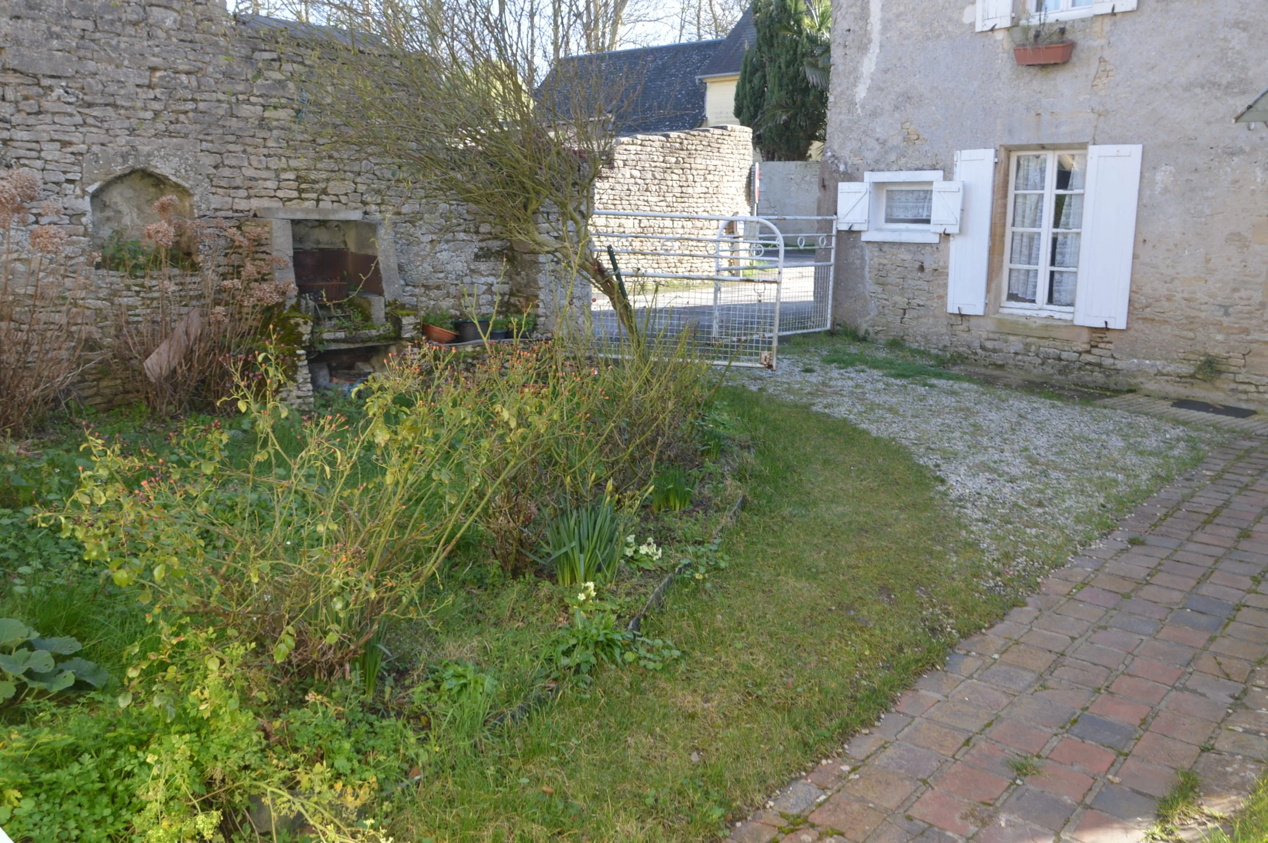 View of a small garden with a stone wall, plants, a gravel patch, a brick pathway, and a stone building with white shutters.