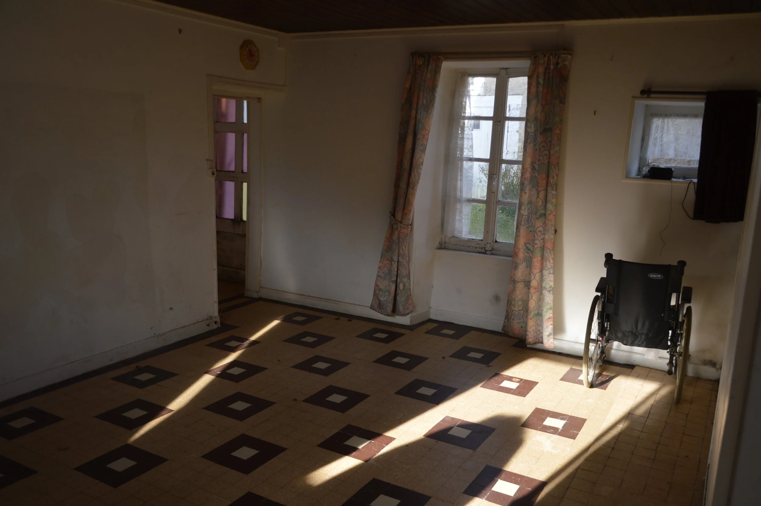 Empty room with patterned linoleum floor, two windows with curtains, and a wheelchair near the wall.