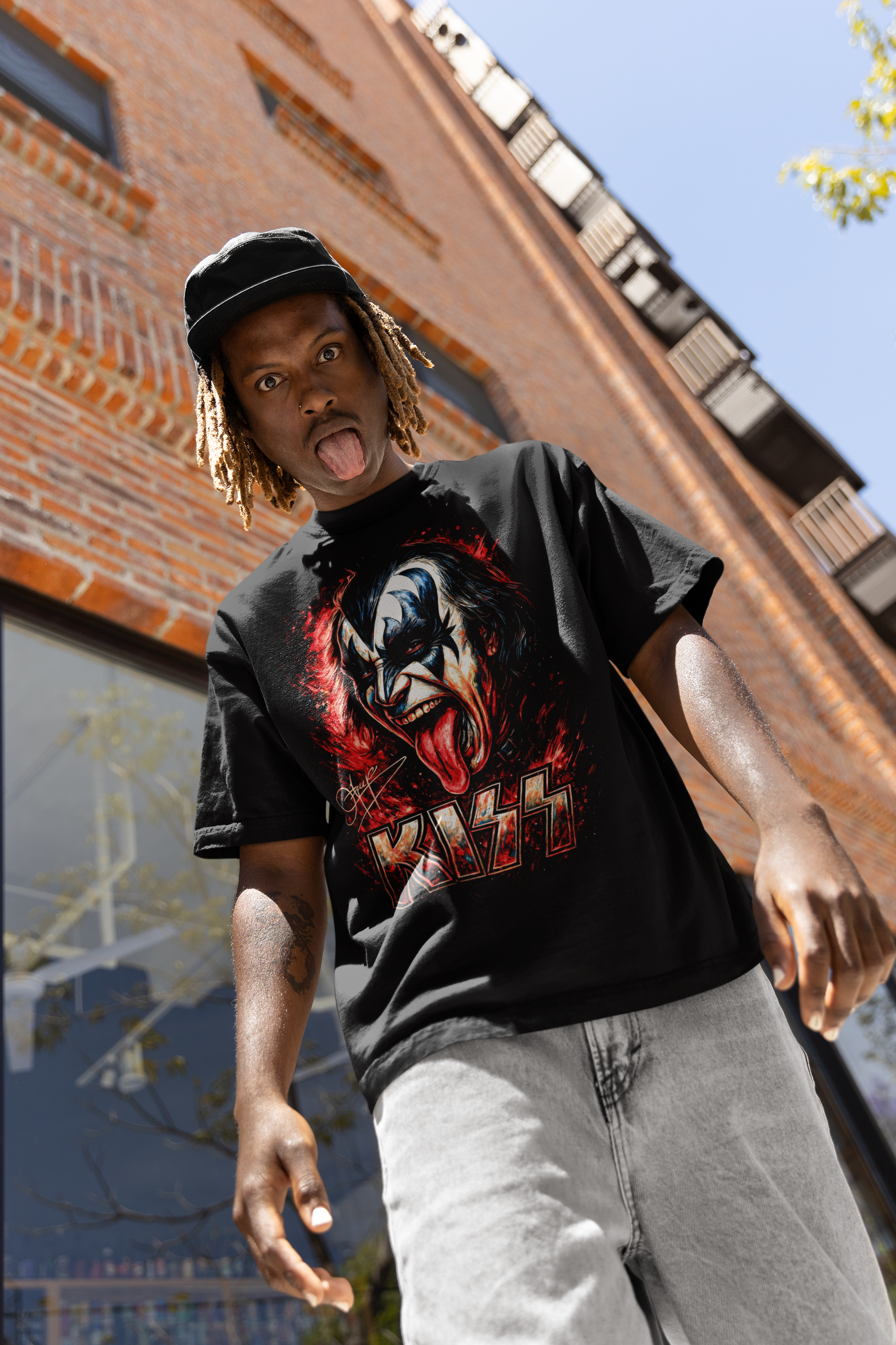 A young man with dreadlocks sticking out of a black cap, wearing a black KISS band T-shirt and light gray jeans, sticking out his tongue and looking down at the camera outside a brick building.