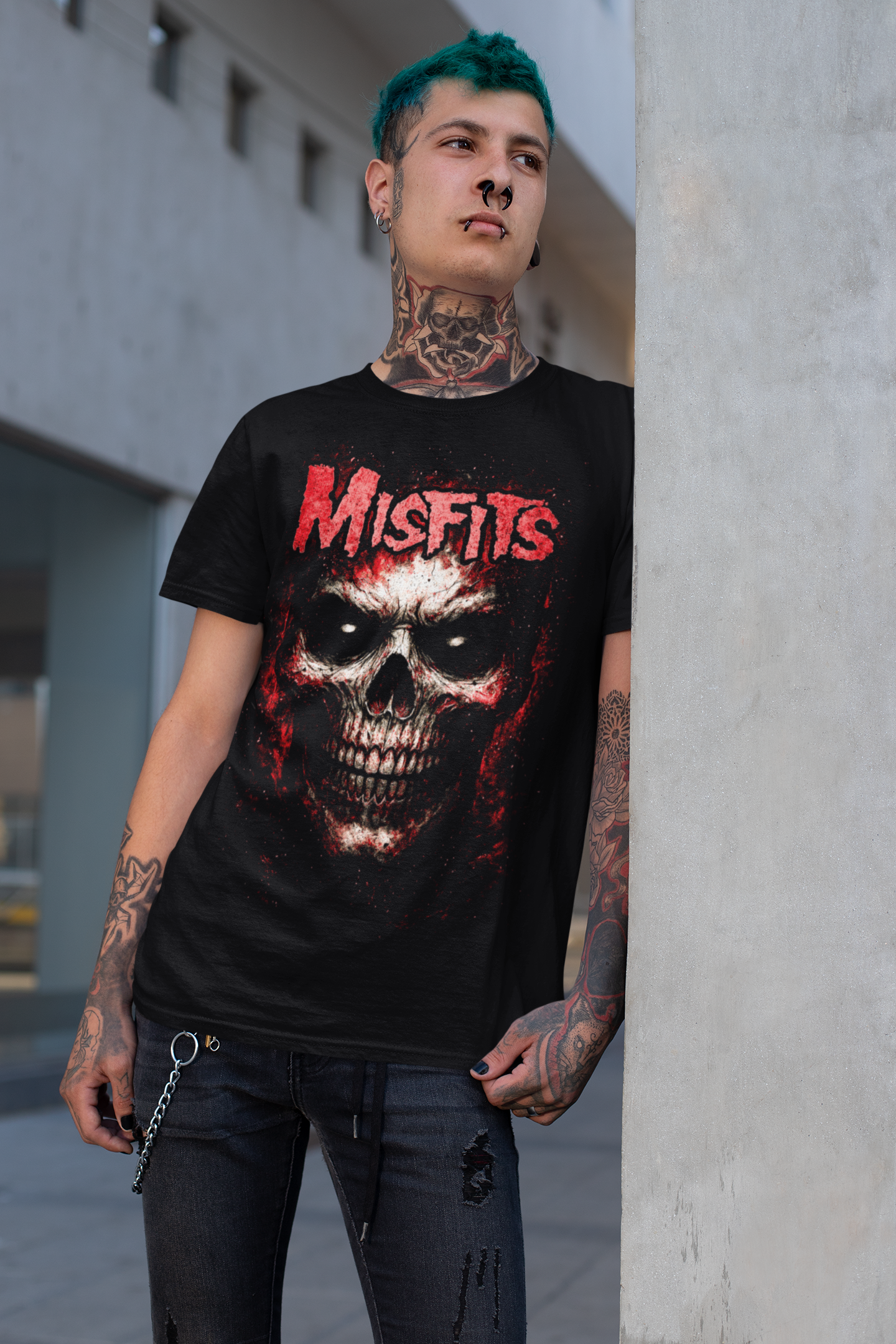 A young man with blue hair, tattoos, and piercings wearing a Misfits band T-shirt with a skull graphic, standing next to a concrete wall in an urban environment.