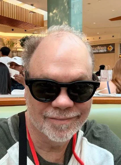 A man with glasses and a beard smiling in a busy indoor setting, possibly a restaurant or shopping mall.