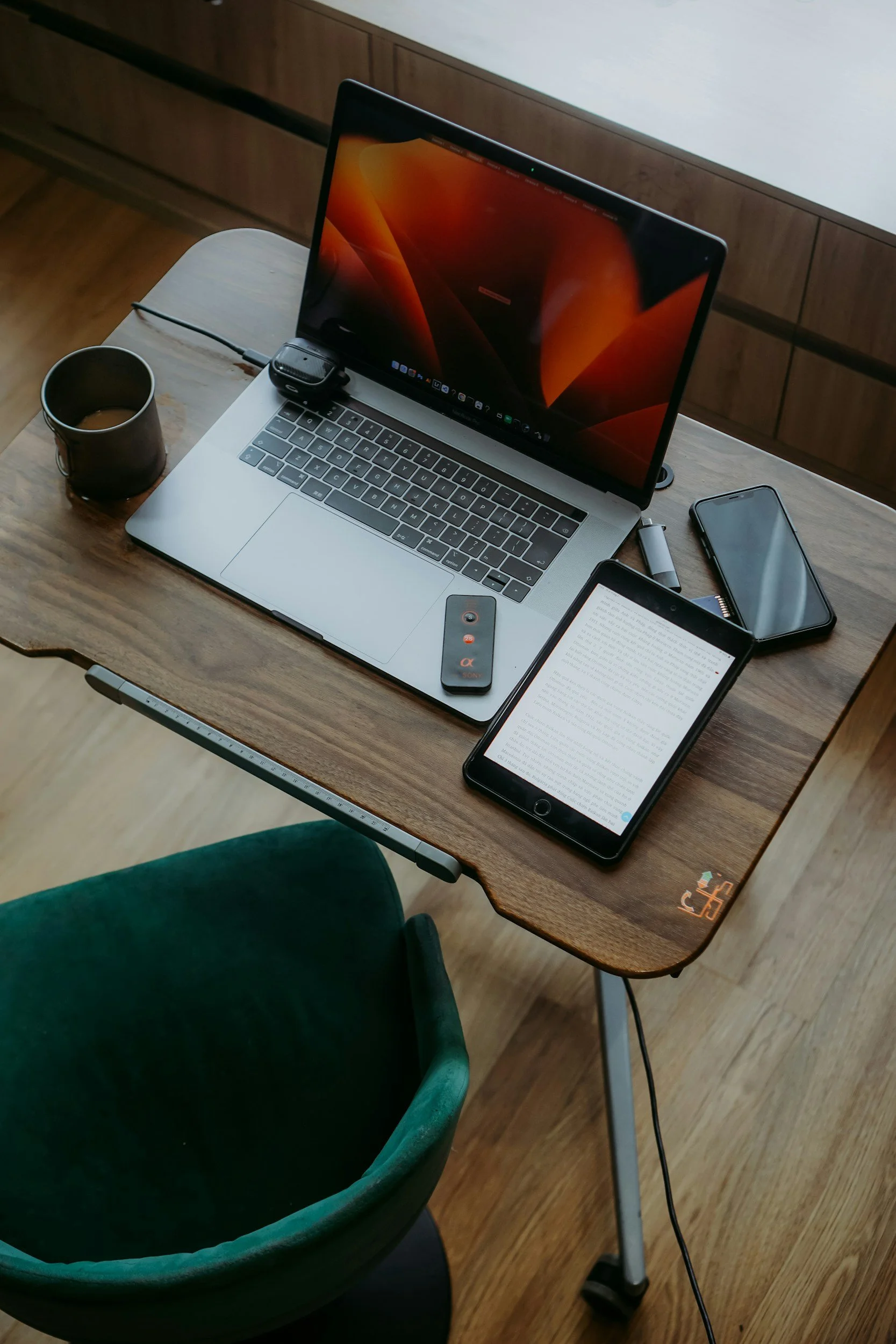 A wooden desk with an open laptop, a smartphone, a tablet with text, a black coffee mug, and various small electronic accessories, next to a green upholstered chair.