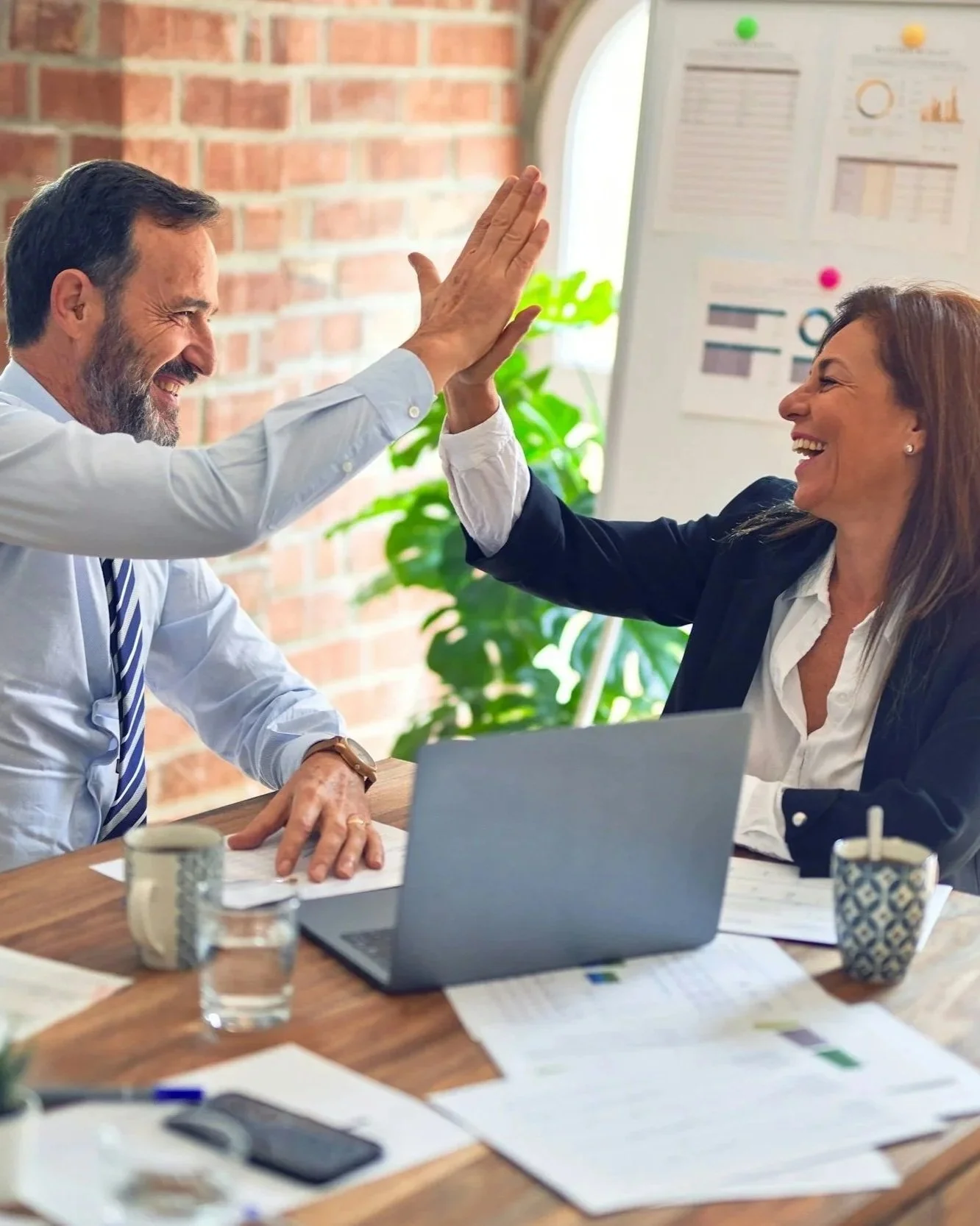 A man and a woman in business attire giving each other a high five in an office setting with documents, coffee mugs, and a laptop on the table.
