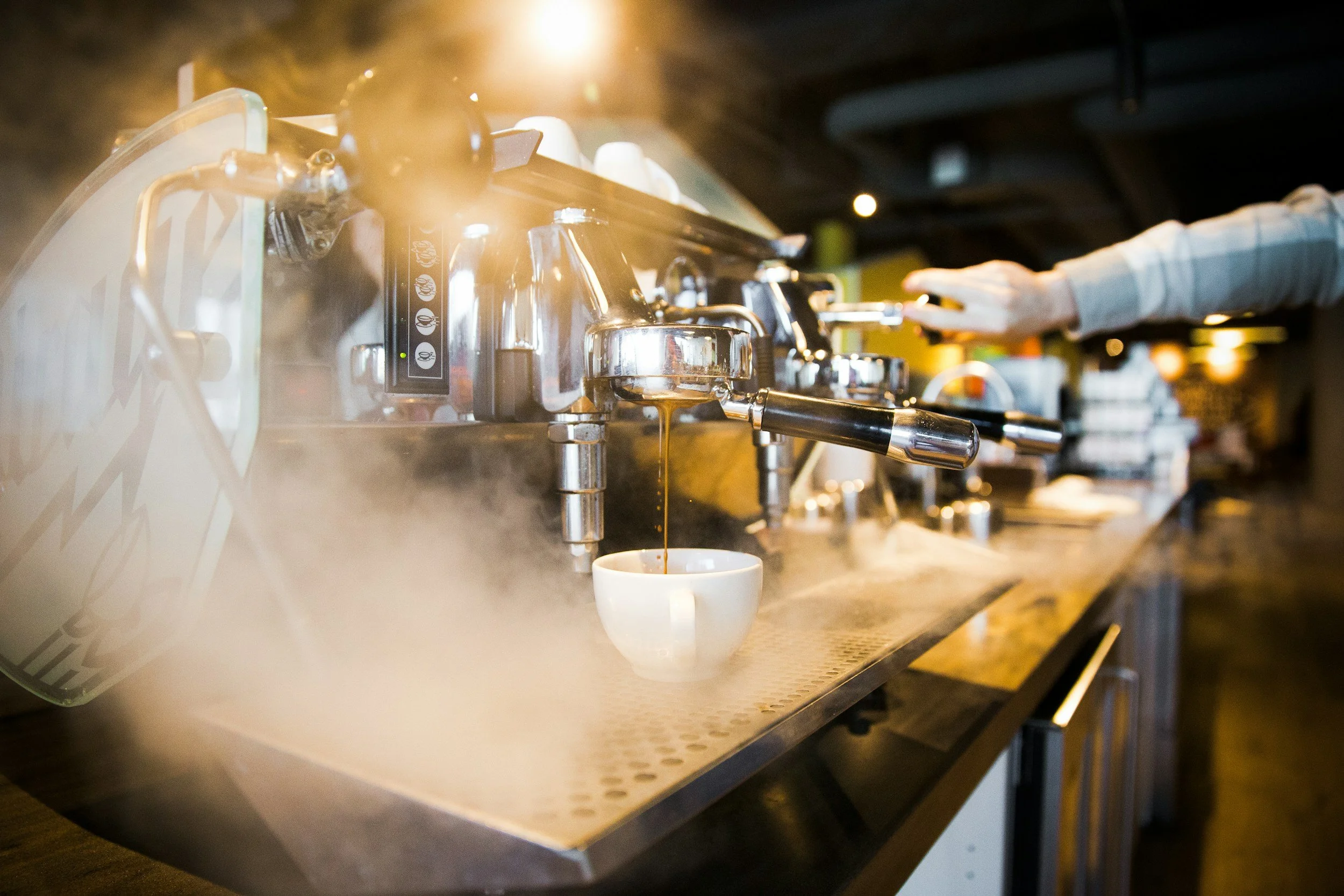 A barista uses a coffee machine to brew espresso into a white cup in a cafe.
