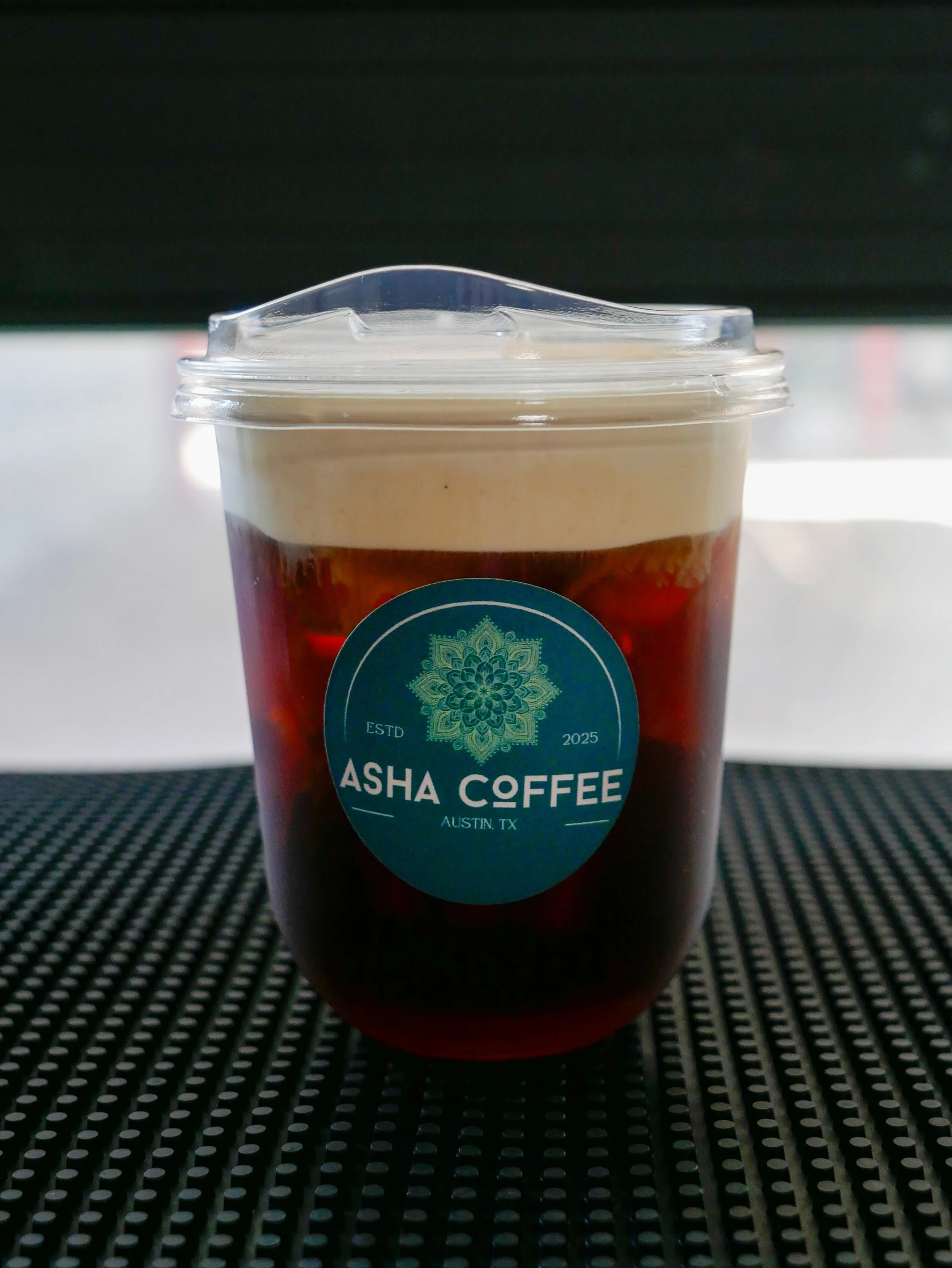 A plastic cup of cold coffee topped with whipped cream, with a blue logo that says 'Asha Coffee, Austin, TX, Estd 2025'.