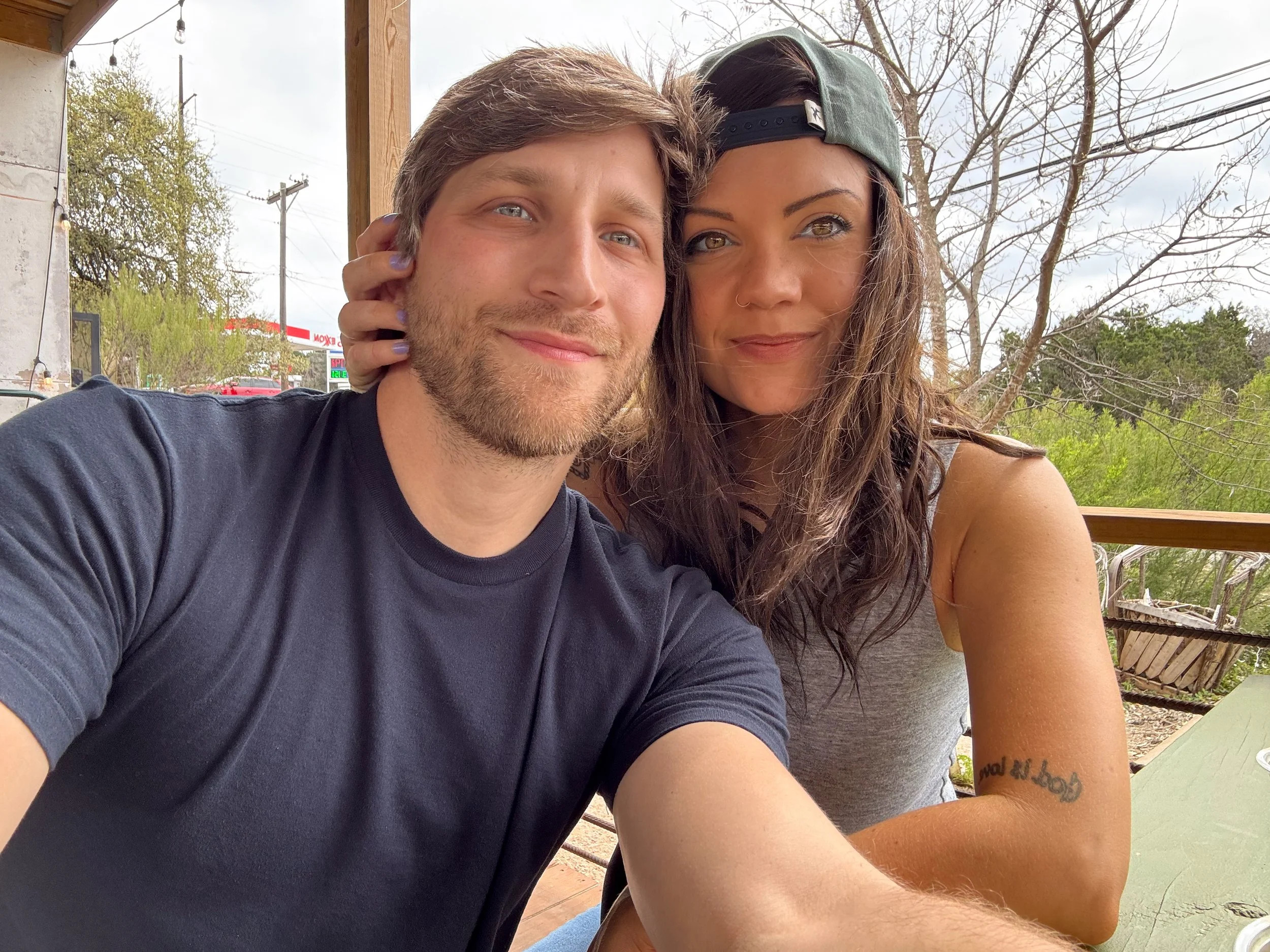 A young man and woman taking a selfie outdoors on a patio. The man has blonde hair and a beard, wearing a navy T-shirt. The woman has brown hair, wearing a gray tank top and a backward baseball cap, with a tattoo on her arm.