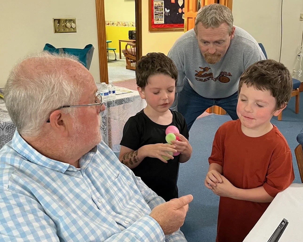 An elderly man with white hair and glasses is seated and looking at two young boys standing in front of him. One boy, wearing a black shirt, is holding a small colorful toy and looking at the elderly man. The other boy, wearing a red shirt, is standing with hands clasped, also watching the elderly man. A man with a beard and gray shirt is kneeling behind the boys, observing the scene, in a room with wooden trim and a table with water bottles.