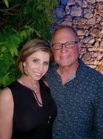 A woman and an older man posing together in front of a stone wall with greenery, smiling at the camera.