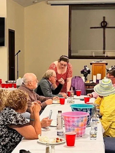 A group of elderly people seated at a table with red cups, bottles of water, and plates, with a woman standing and talking to them, and a Cross on the wall in the background.