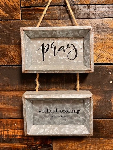 Two rustic metal-framed signs hanging on a wooden wall. The top sign reads "pray" in black script, and the bottom sign features a Bible verse, 1 Thessalonians 5:17, "without ceasing," in black text.