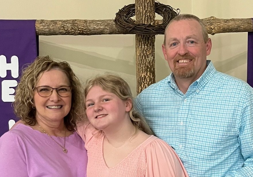 A family of three standing together indoors in front of a wooden cross. The woman on the left is wearing glasses and a purple top, the girl in the middle is wearing a pink dress, and the man on the right is wearing a blue checkered shirt. They are smiling and posing for the photo.