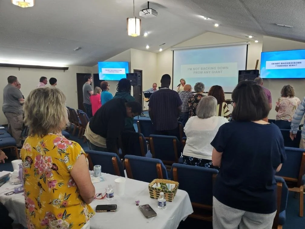 People gathered in a church or conference room, standing and facing a stage with a large screen displaying lyrics, during a worship or prayer event.