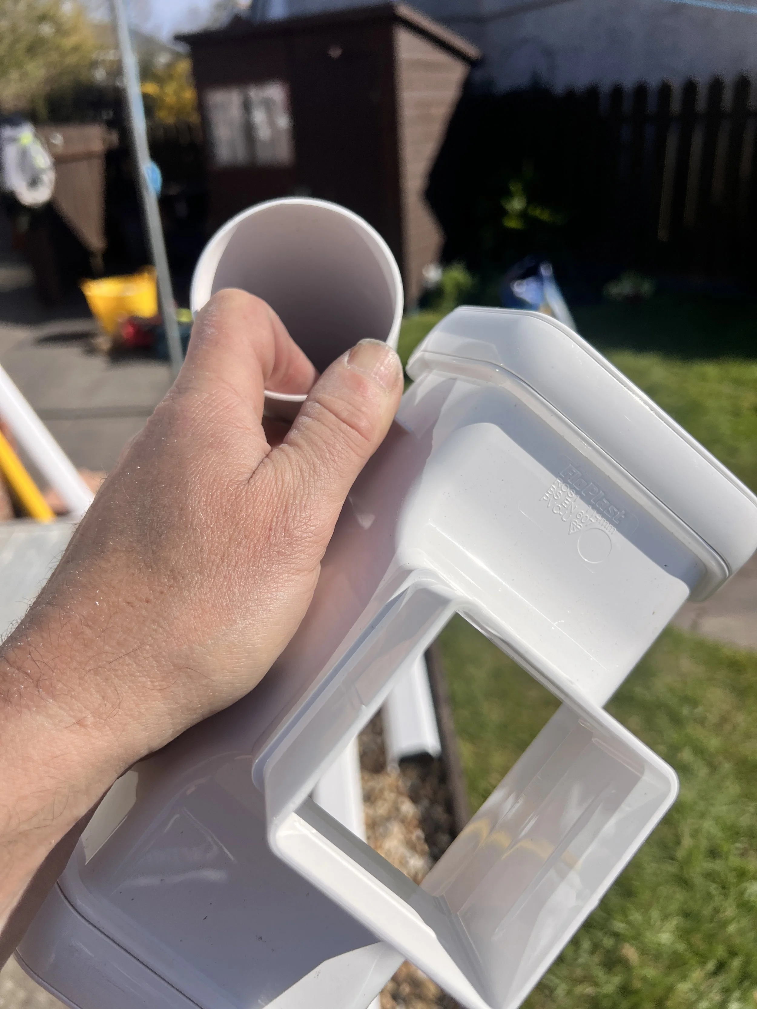A person's hand holding a white plastic container or extension of a sprayer, with a blurred background of a brown shed, a blue sky, and a grassy yard.
