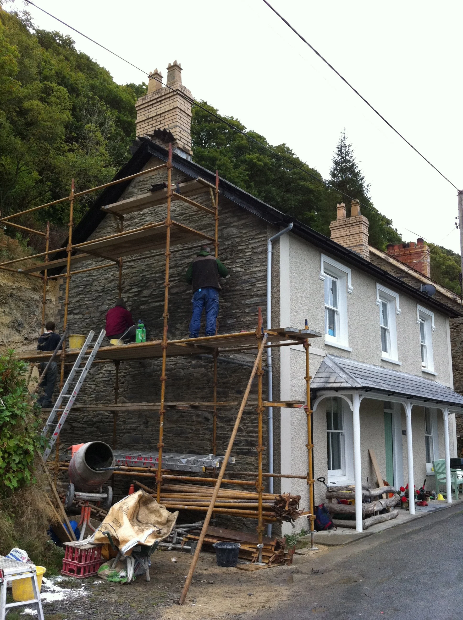 Construction workers working on a house with scaffolding, replacing the exterior wall with stone veneer. Tools, materials, and a cement mixer are present in the yard.