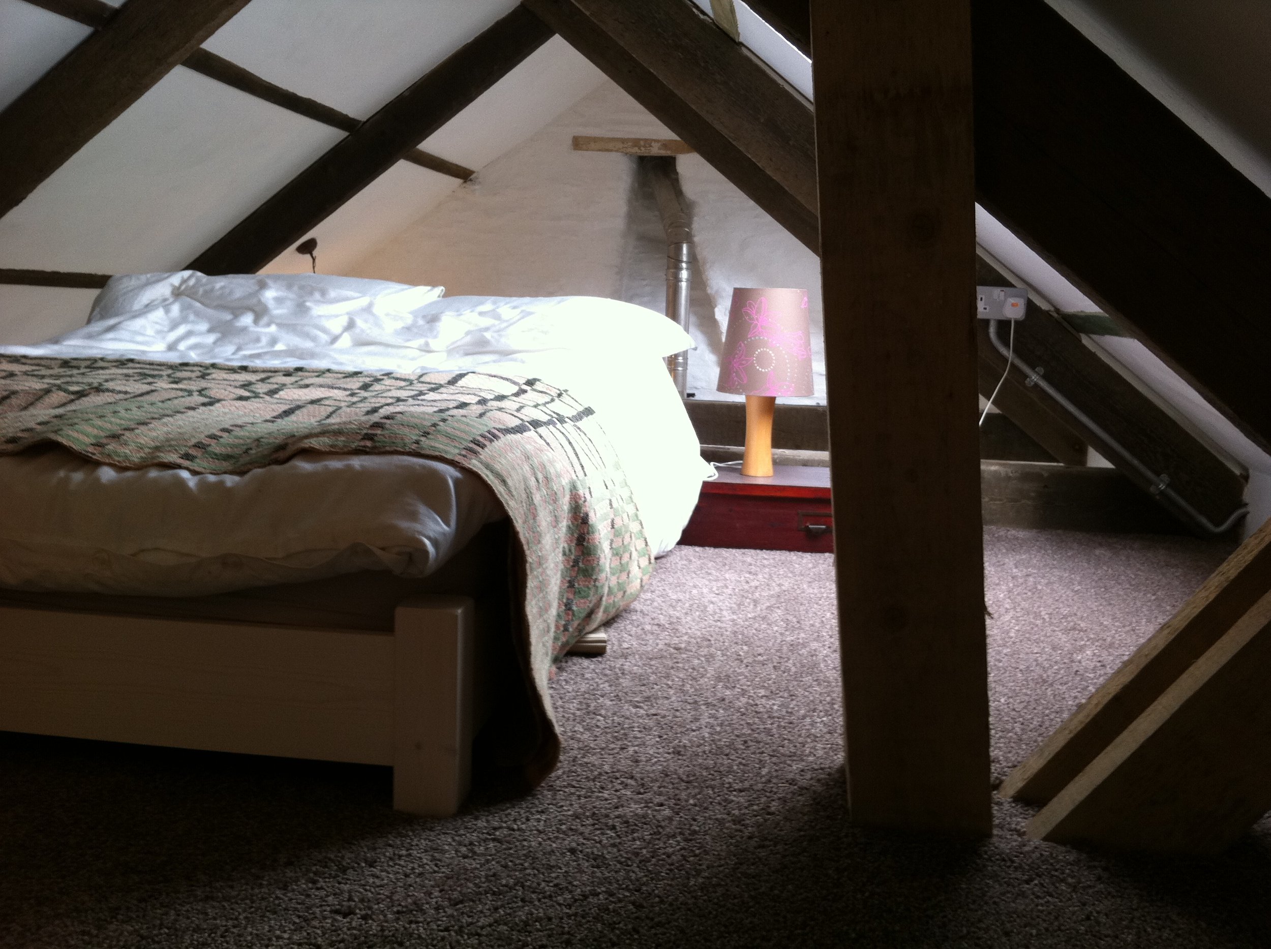 A bedroom with a bed covered in white and a blanket. A pink lamp with flower pattern on a nightstand next to the bed. The room has sloped ceilings with dark wooden beams and a carpeted floor.