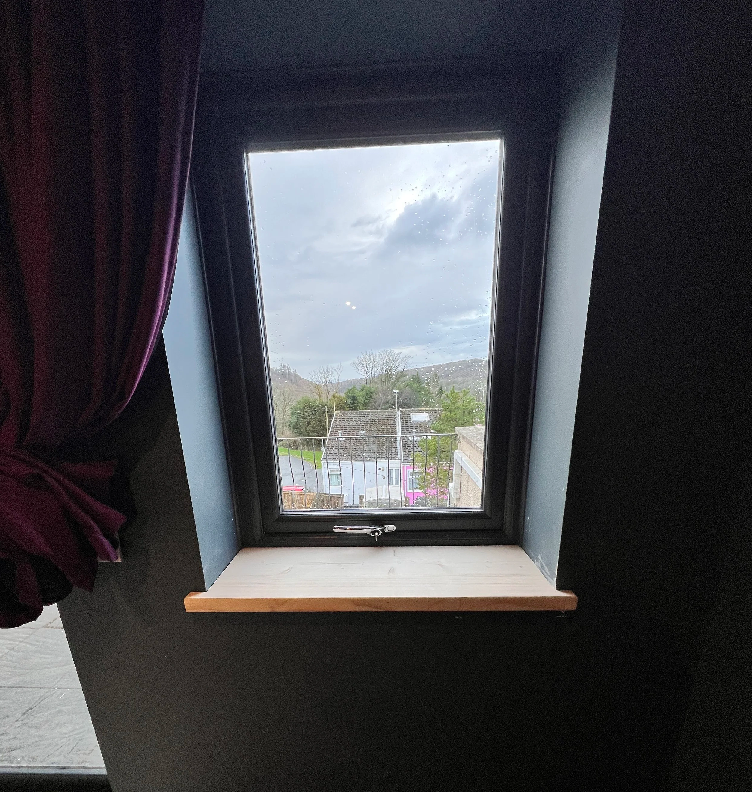 A view through a rectangular window showing a cloudy sky, some hills, trees, rooftops, and a street below. The window has dark framing and purple curtains on the side.