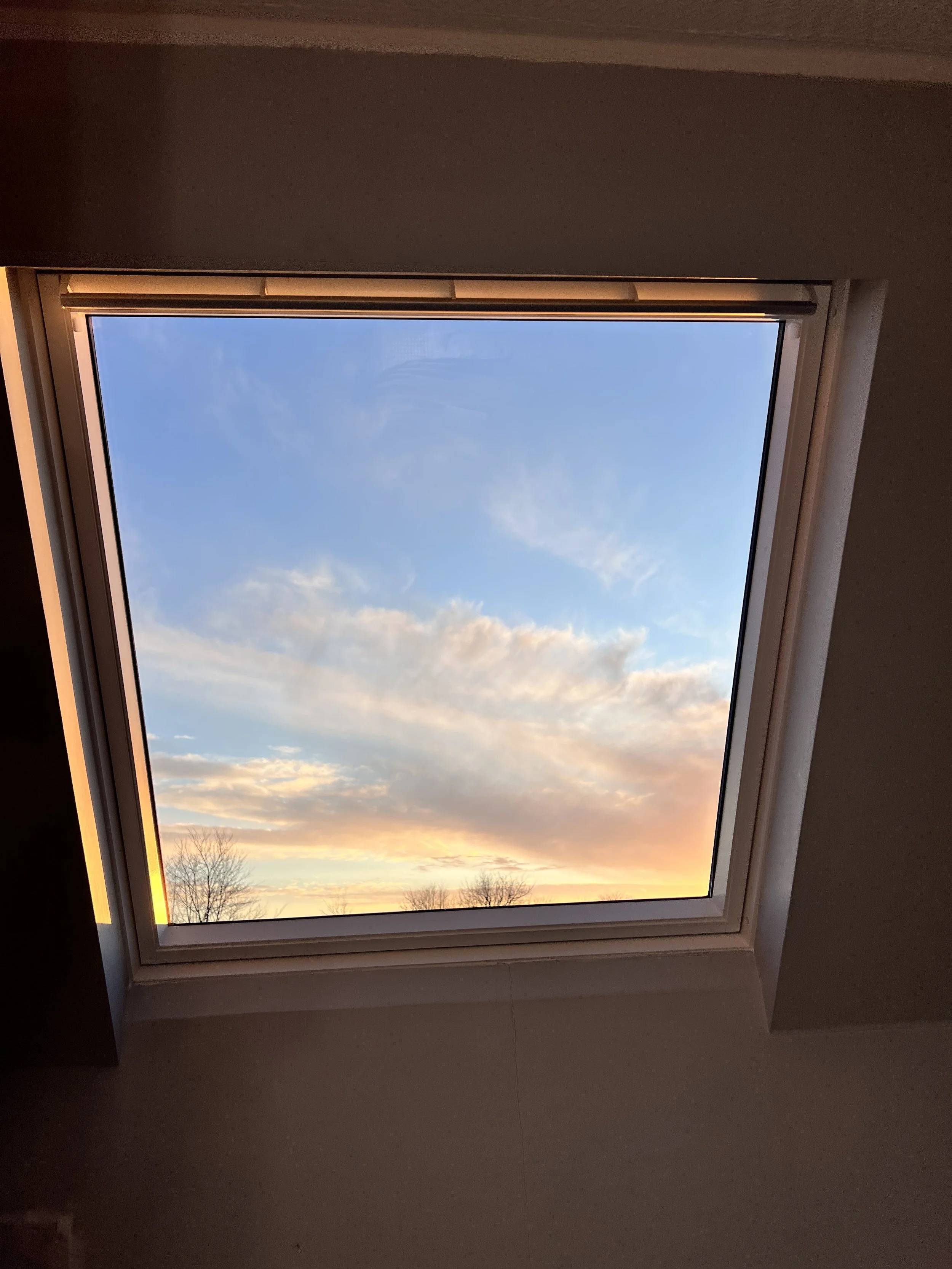 View of the sky through a rectangular skylight during sunset, with clouds and bare tree silhouettes visible.