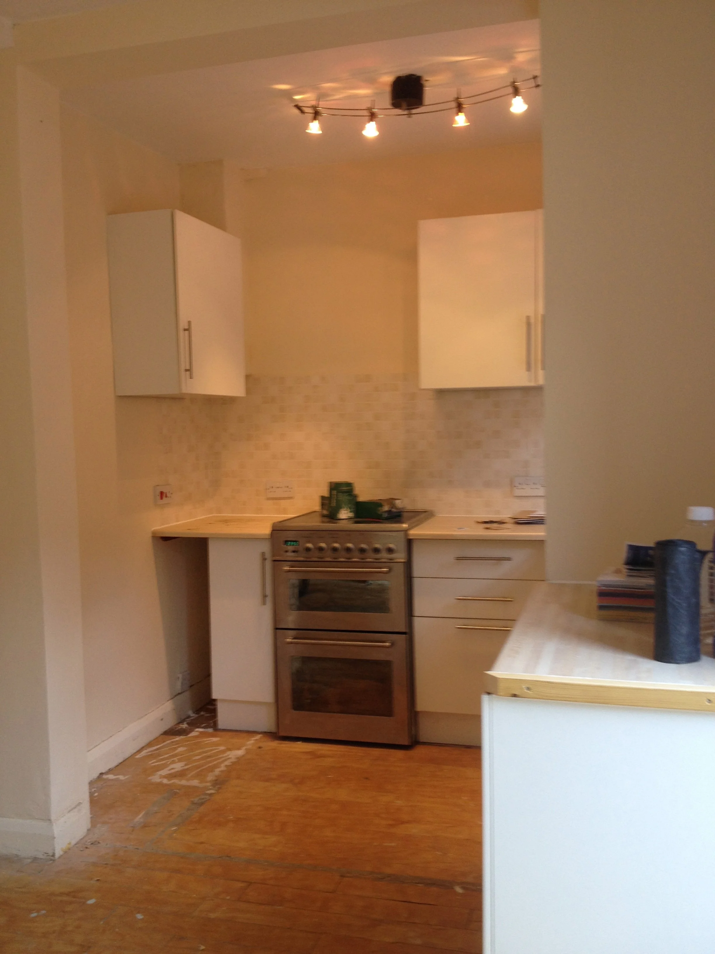 A kitchen with white cabinetry, a stove, and a wooden countertop, with a peeled wooden floor and some boxes and items on the counter.