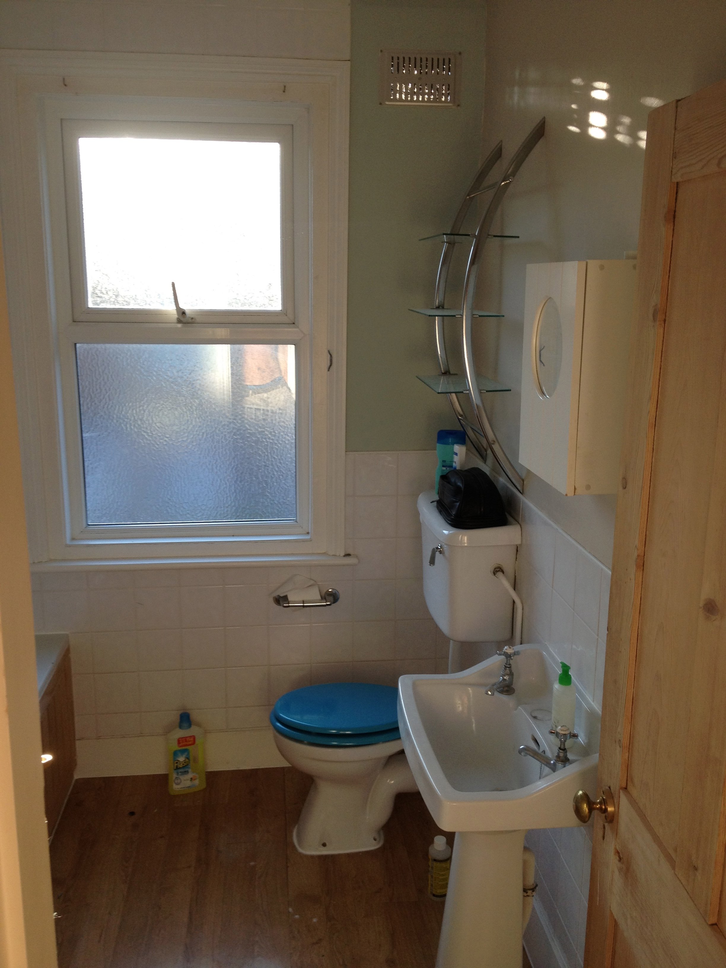 A small bathroom featuring a window with frosted glass, a toilet, a bidet, a wooden cabinet, a set of glass shelves, a bottle of cleaning solution, a box of tissues, and a soap dispenser on the sink.