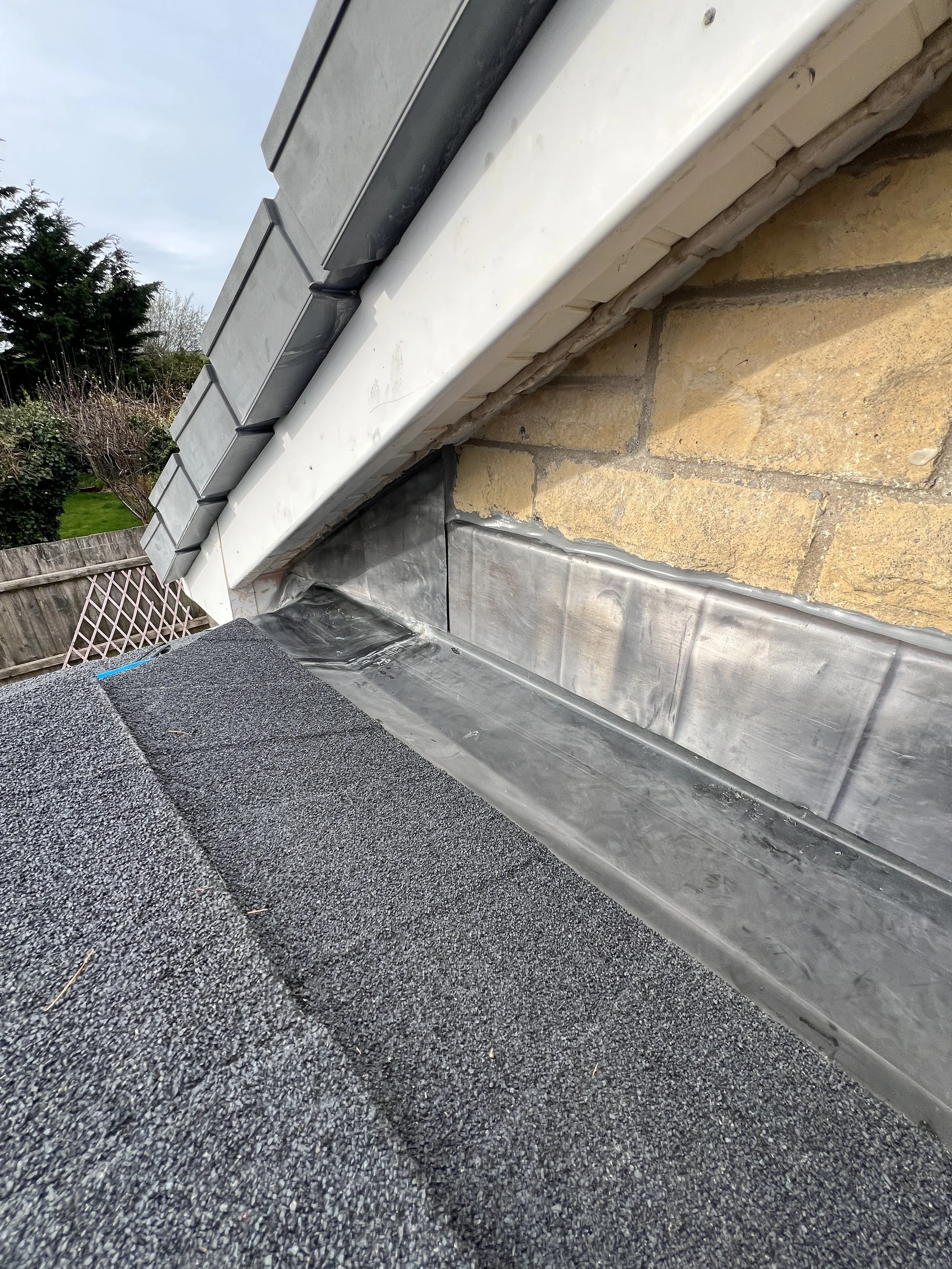Close-up view of a roof gutter system with flashing, fascia, and shingles, alongside bricks and concrete on a building exterior.