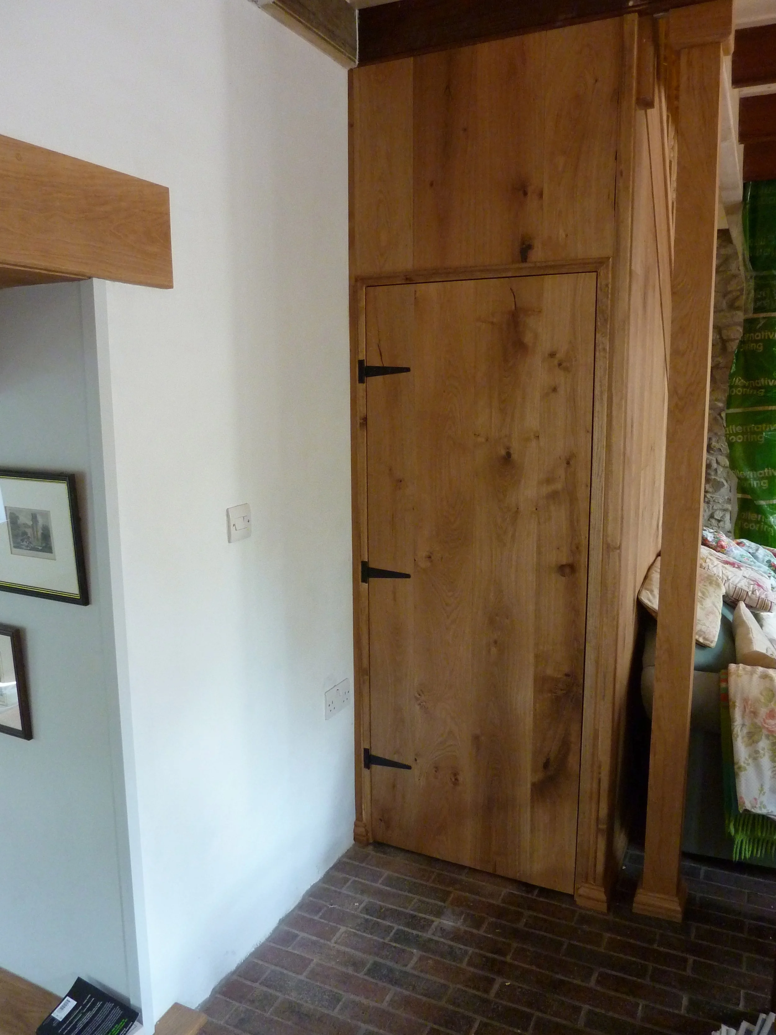 A large wooden cabinet with doors and black metal hinges and handles, standing on a brick floor next to a white wall in a room with framed pictures on the wall.