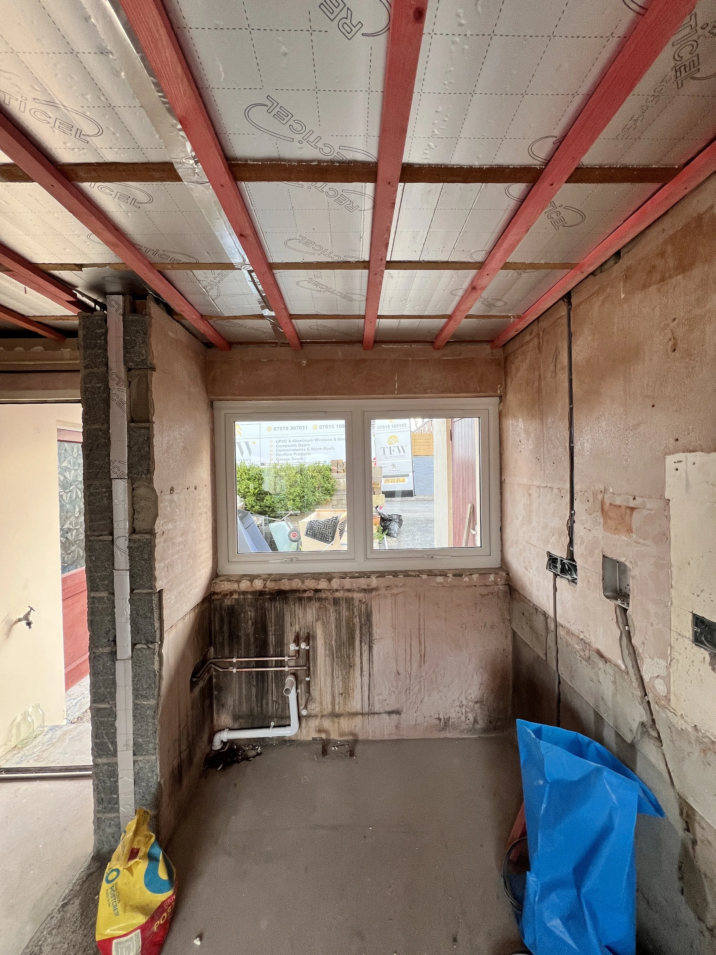 Interior of a room under renovation with exposed wall studs, electrical wiring, and plumbing. There is a window captioned with advertising for UPVC and aluminum windows and doors. Construction materials and tools are scattered on the floor.
