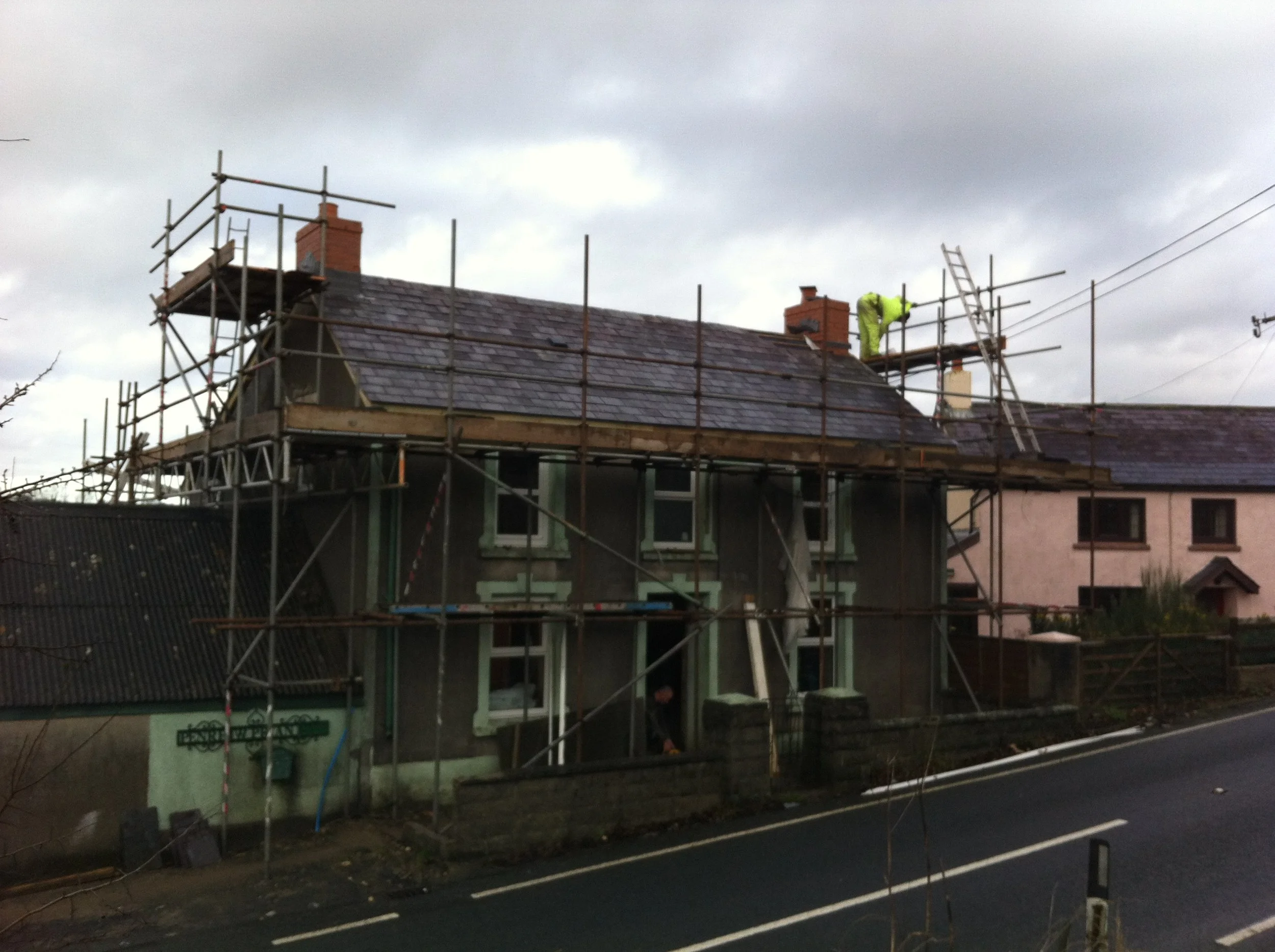 A house under renovation with scaffolding around it, a worker in high-visibility clothing on the roof, and cloudy skies overhead.