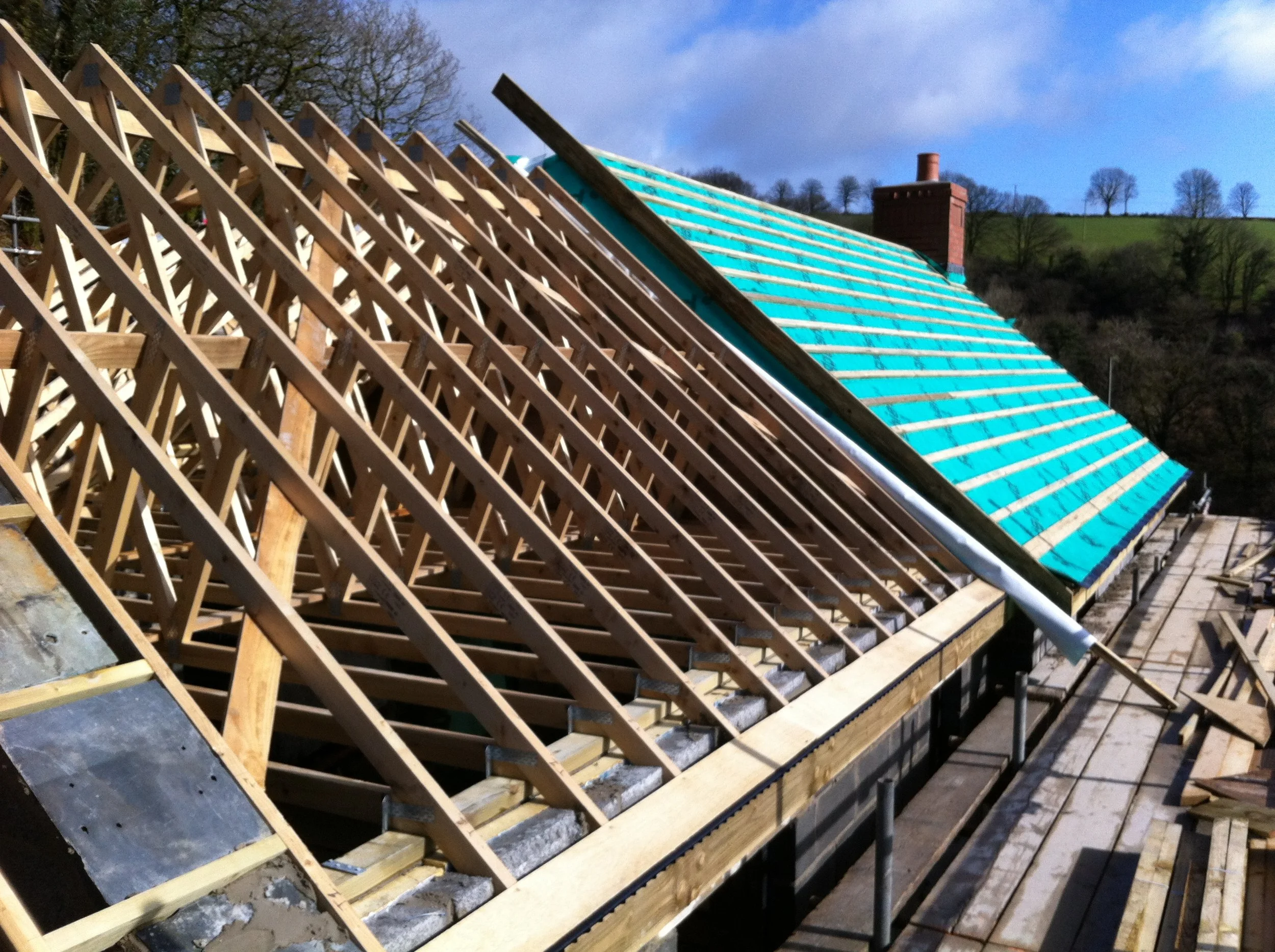 Under construction roof with wooden framework and turquoise tiles, overlooking a green hilly landscape with leafless trees.